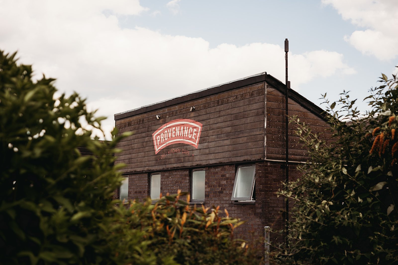 A two-story brick building with wooden siding on the top floor, with a red and white sign reading 'Provenance' and 'Del Kitchen Cafe' beneath it, surrounded by green bushes and trees under a cloudy sky.