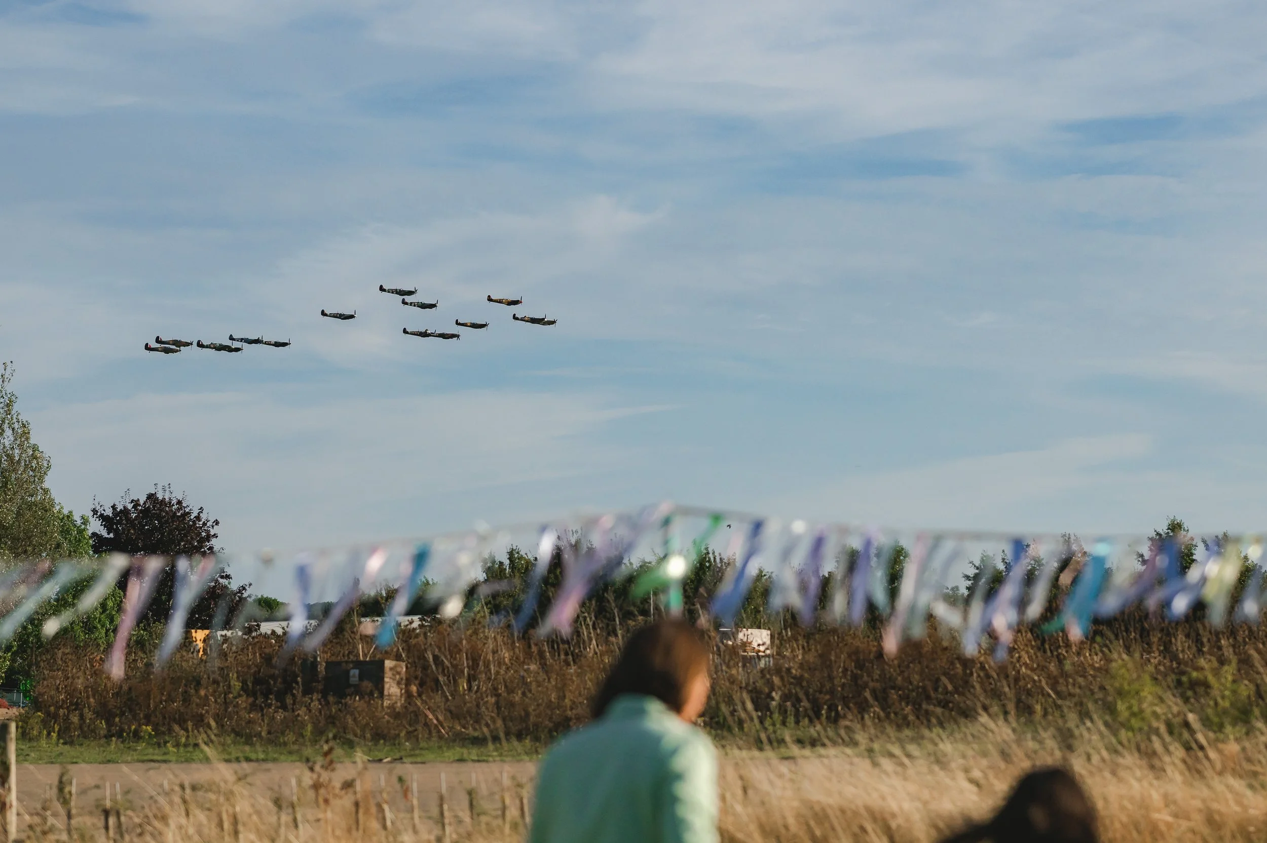 A formation of vintage military aircraft flying in a V-shape in the sky during an air show. In the foreground, there are blurred people and colorful ribbon decorations hanging across the scene, with trees and a field below.