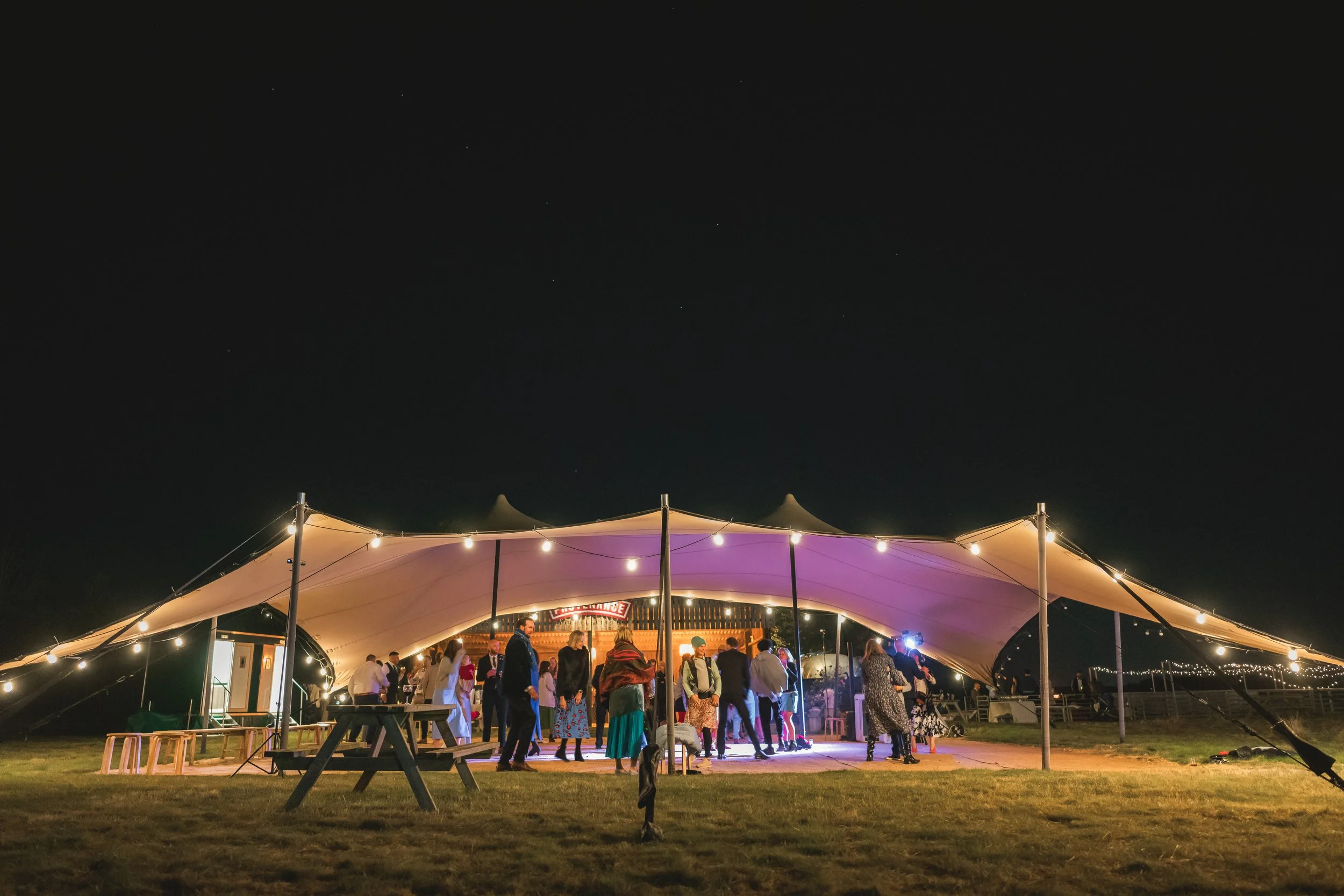 People dancing and socializing under a large outdoor tent with string lights at night.