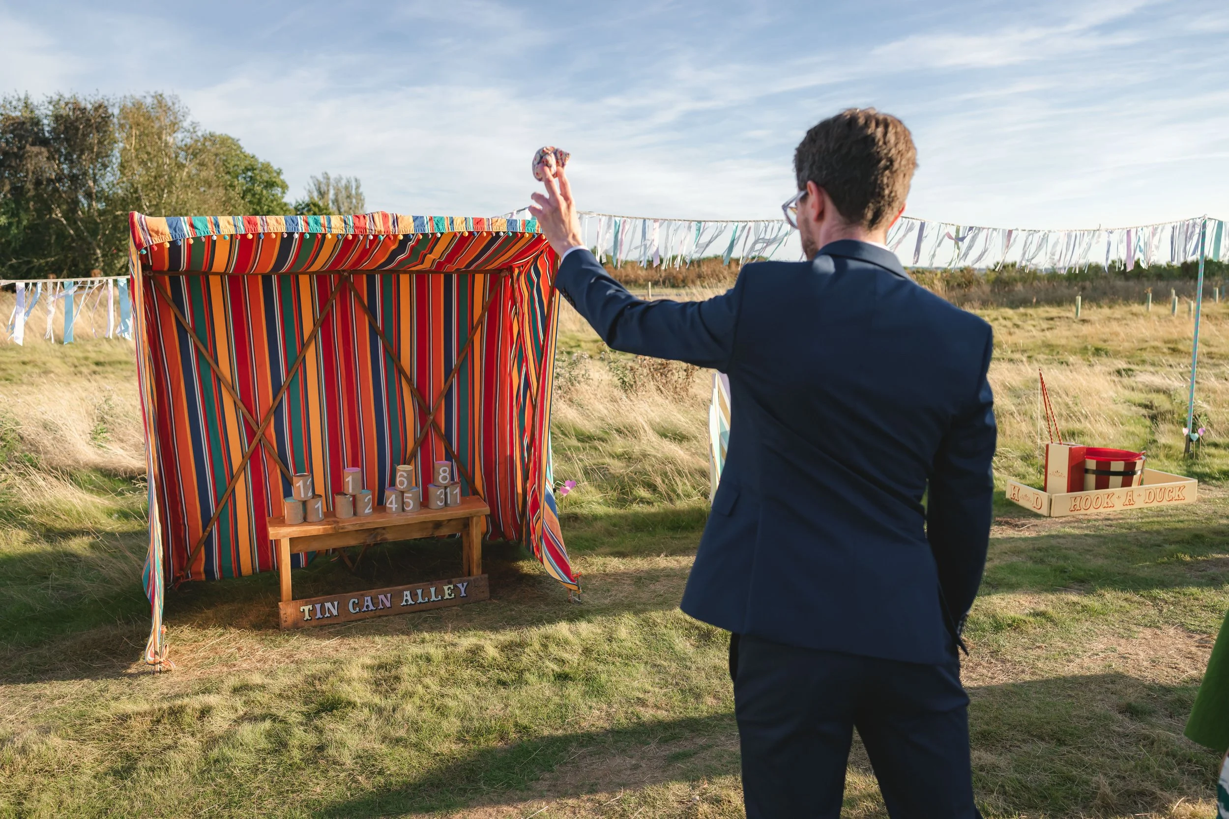 A man in a navy suit throwing a ring at a carnival game called 'Tin Can Alley' with tin cans numbered 1 to 8 stacked on a small wooden bench, set outdoors on a grassy field with a decorative colorful striped tent.