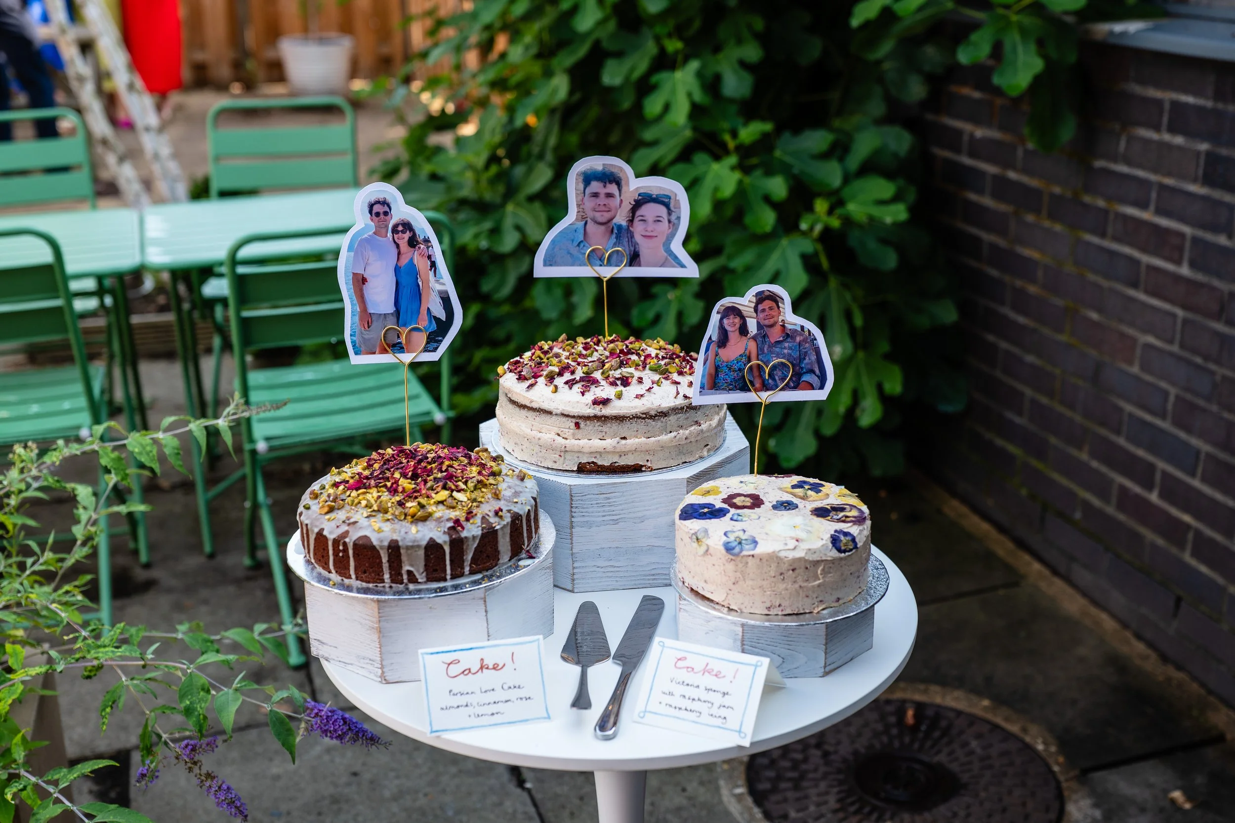 Three decorated cakes with photos of people on sticks on top, set on a white table outdoors with green chairs and foliage in the background.
