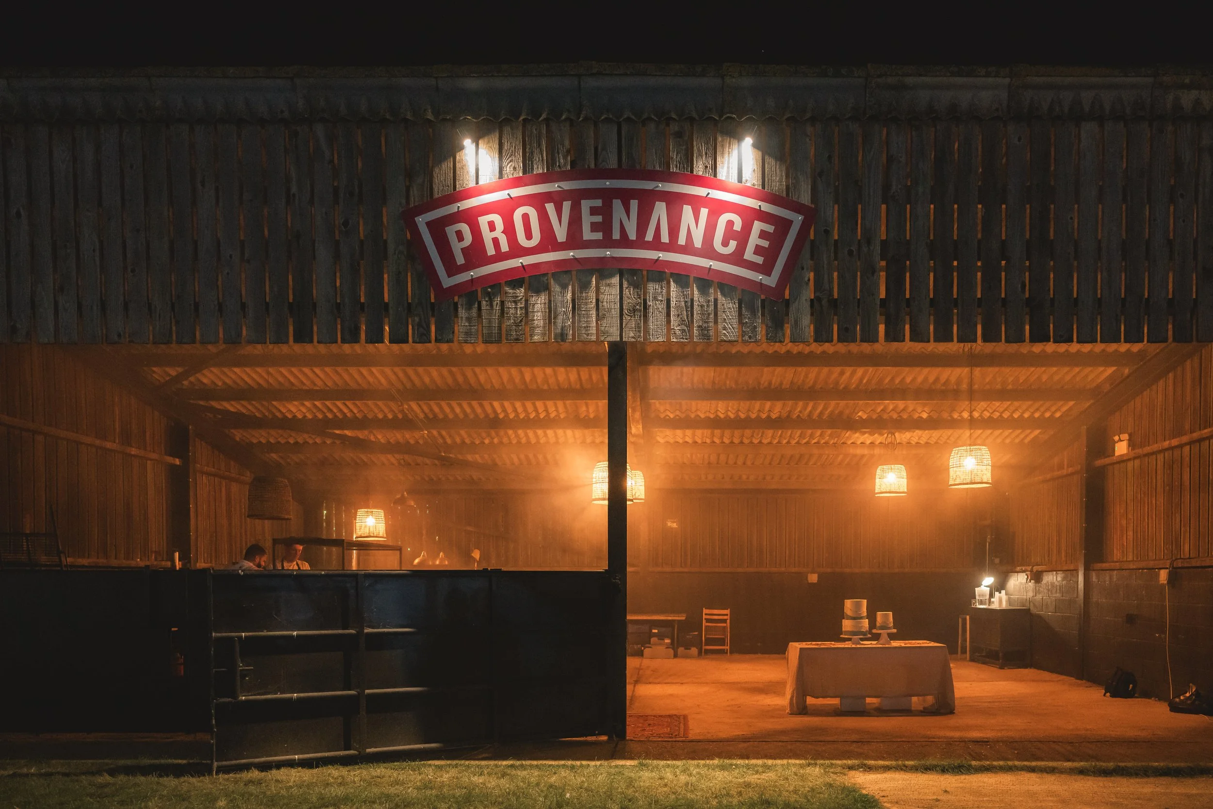 Interior of a rustic event space with wooden walls and ceiling, illuminated by hanging lamps, featuring a sign that says 'Provenance', a table with chairs and decorative items, at night.