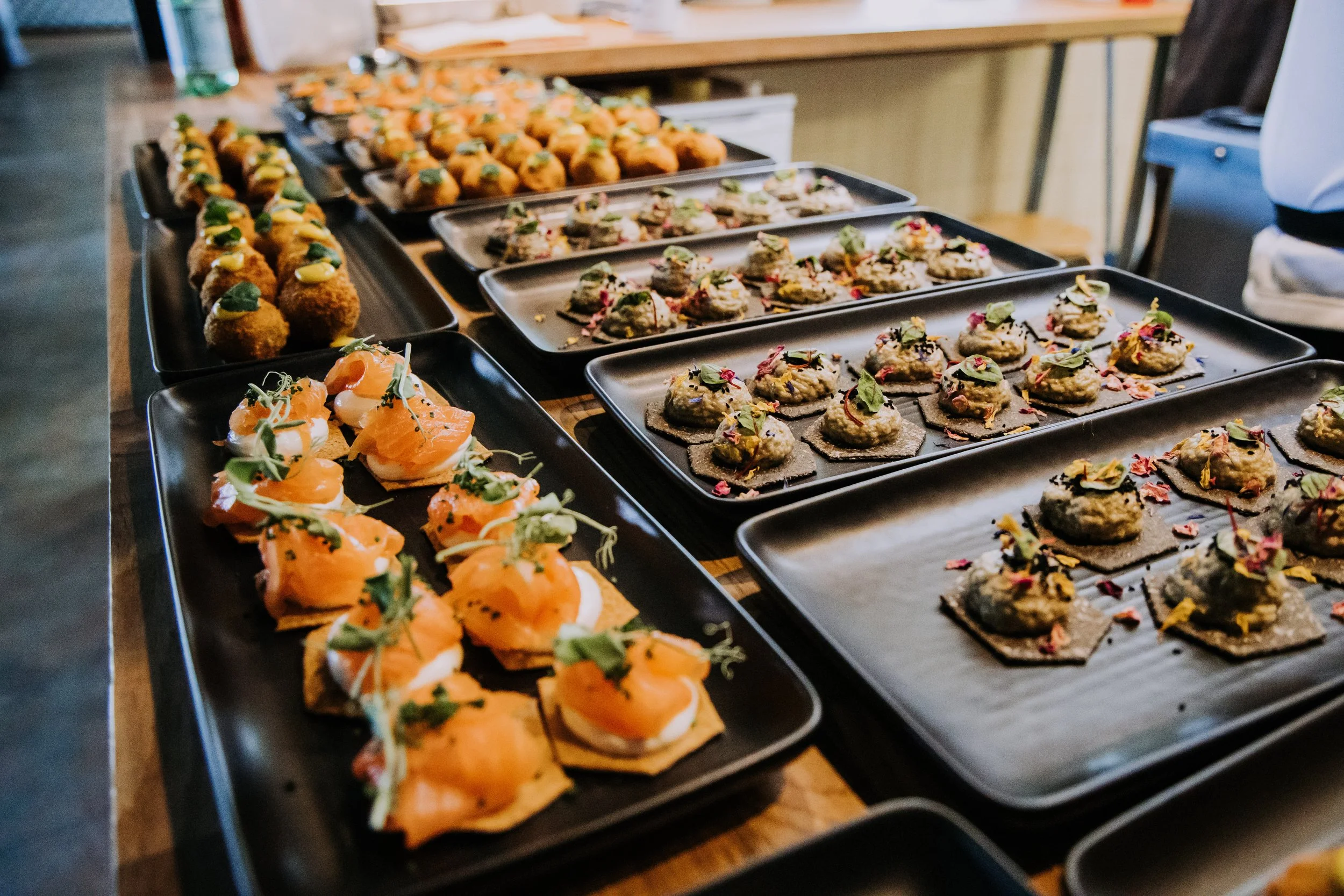 Assorted appetizers on black platters, including smoked salmon canapés, small breaded fritters, and savory bites garnished with herbs, served on a wooden table.