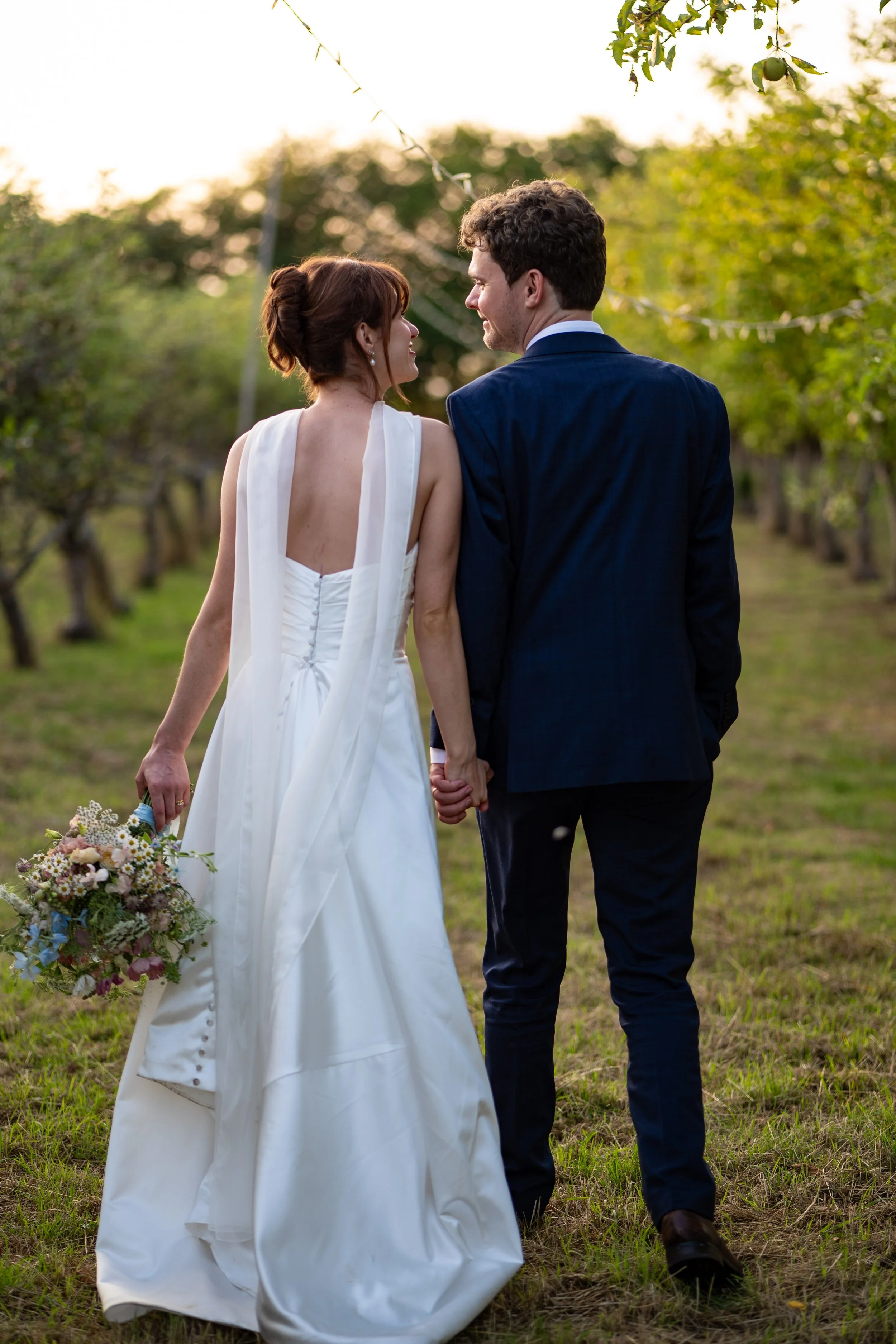 A bride and groom walking hand in hand through a grassy outdoors area, surrounded by trees and string lights, during sunset.