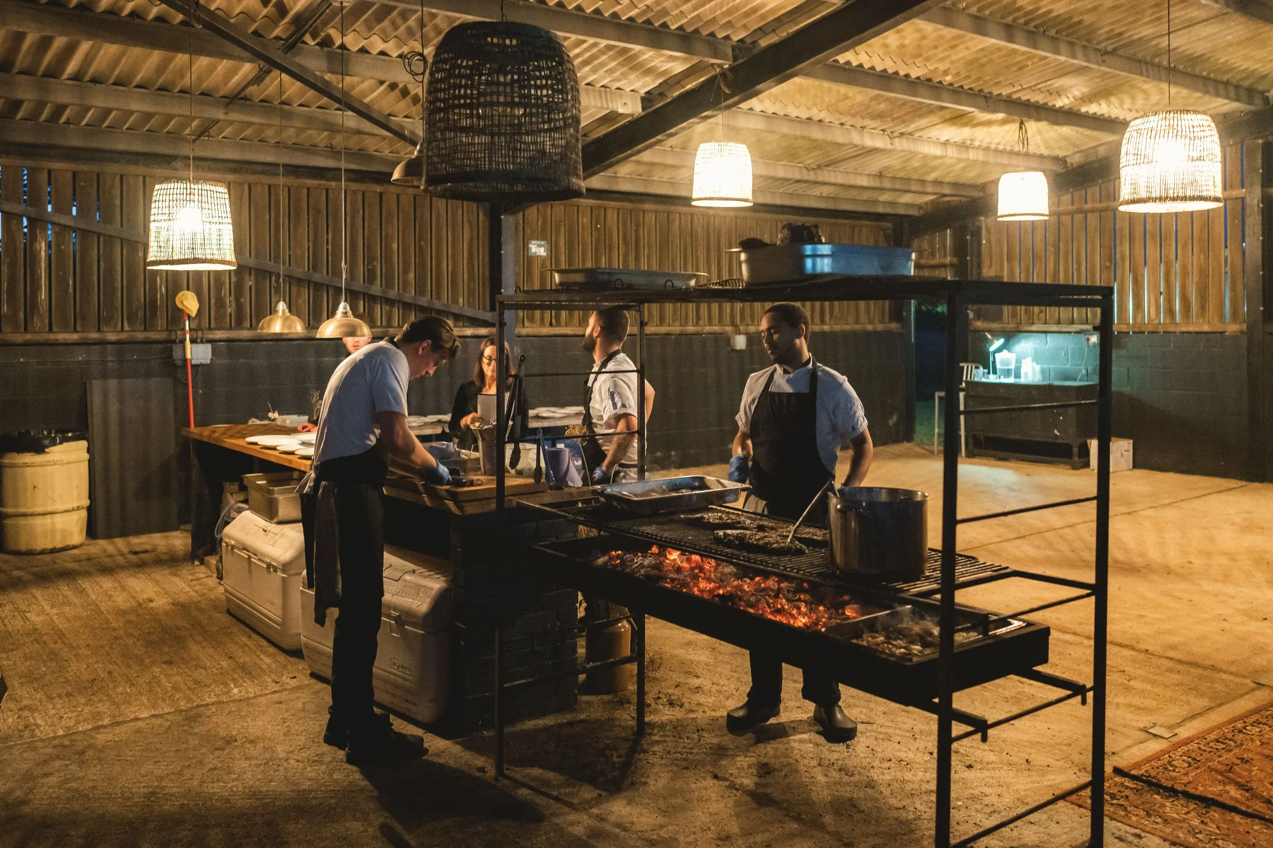 Chefs cooking outdoors in a rustic open kitchen with hanging lamps and warm lighting.