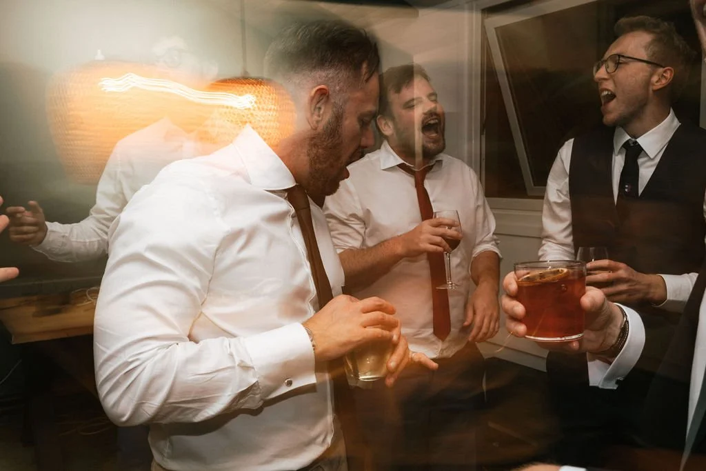 A group of men in formal attire, laughing and drinking cocktails at a party.