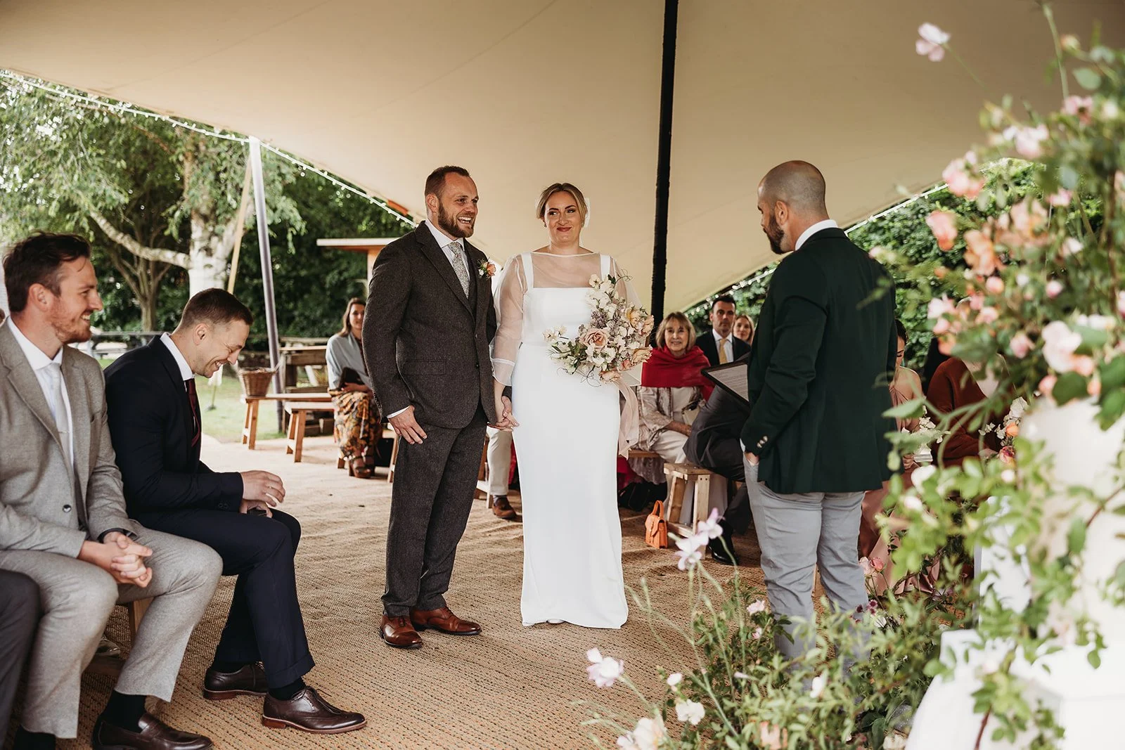 A wedding ceremony under a large canopy with a bride and groom holding hands facing a man officiating. Guests seated around are smiling and watching the couple. The bride wears a white dress and holds a bouquet, while the groom wears a gray suit.