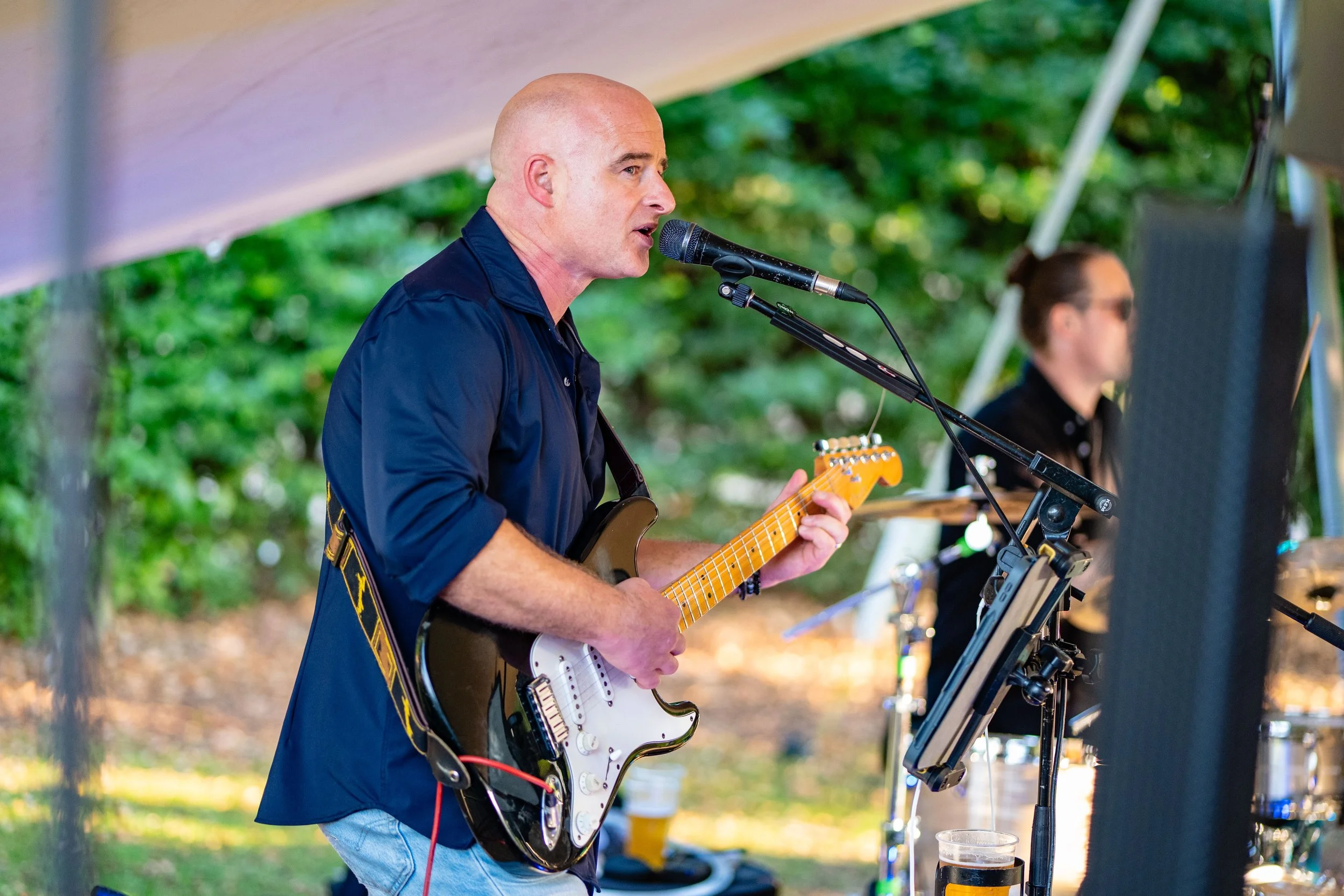 A man playing an electric guitar and singing into a microphone outdoors, with a woman on drums in the background.