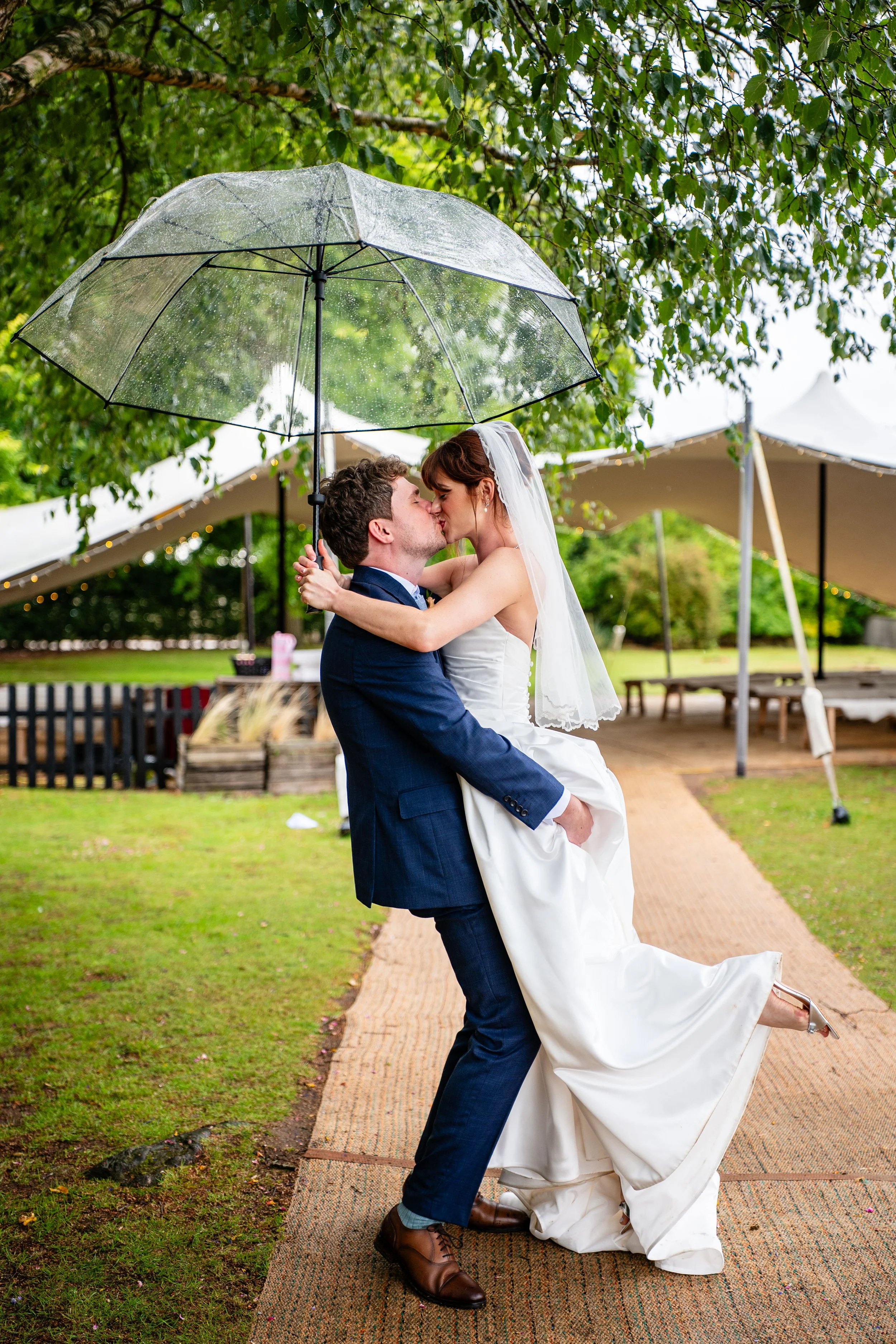 A newlywed couple sharing a kiss under a transparent umbrella outdoors on a rainy day, with green trees and outdoor tent in the background.