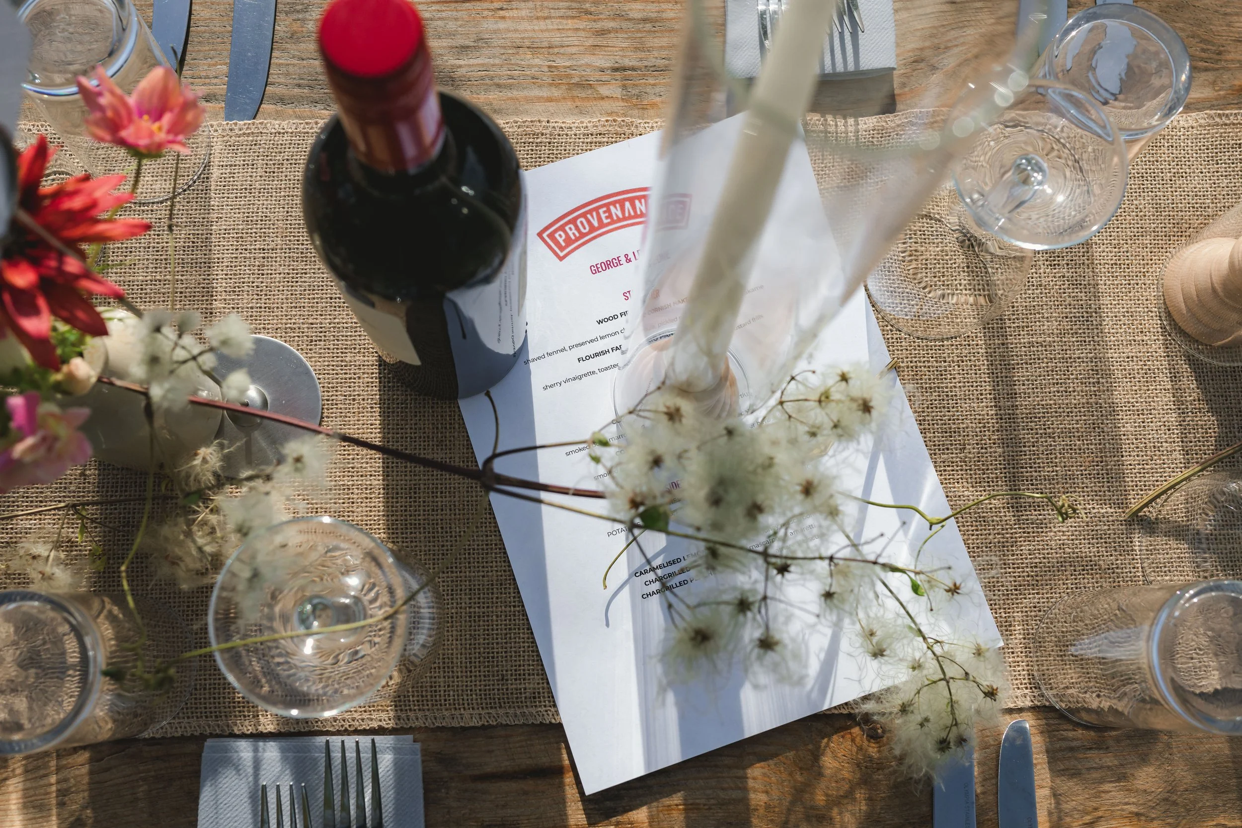 Overhead view of a rustic dining table with a paper menu labeled 'Provenance,' a bottle of red wine, clear glasses, a flower arrangement with pink and white flowers, and a set of tableware on a burlap tablecloth.