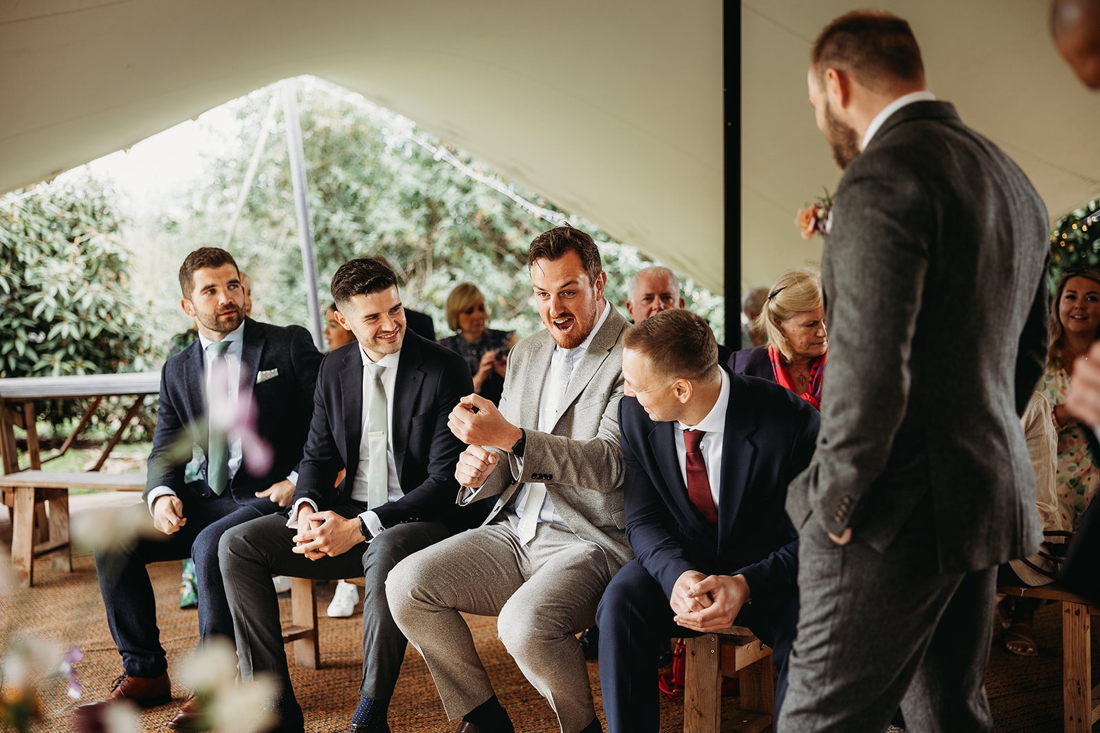 A group of men dressed in suits seated under a tent at a wedding, with one man smiling and gesturing as he talks to another man.