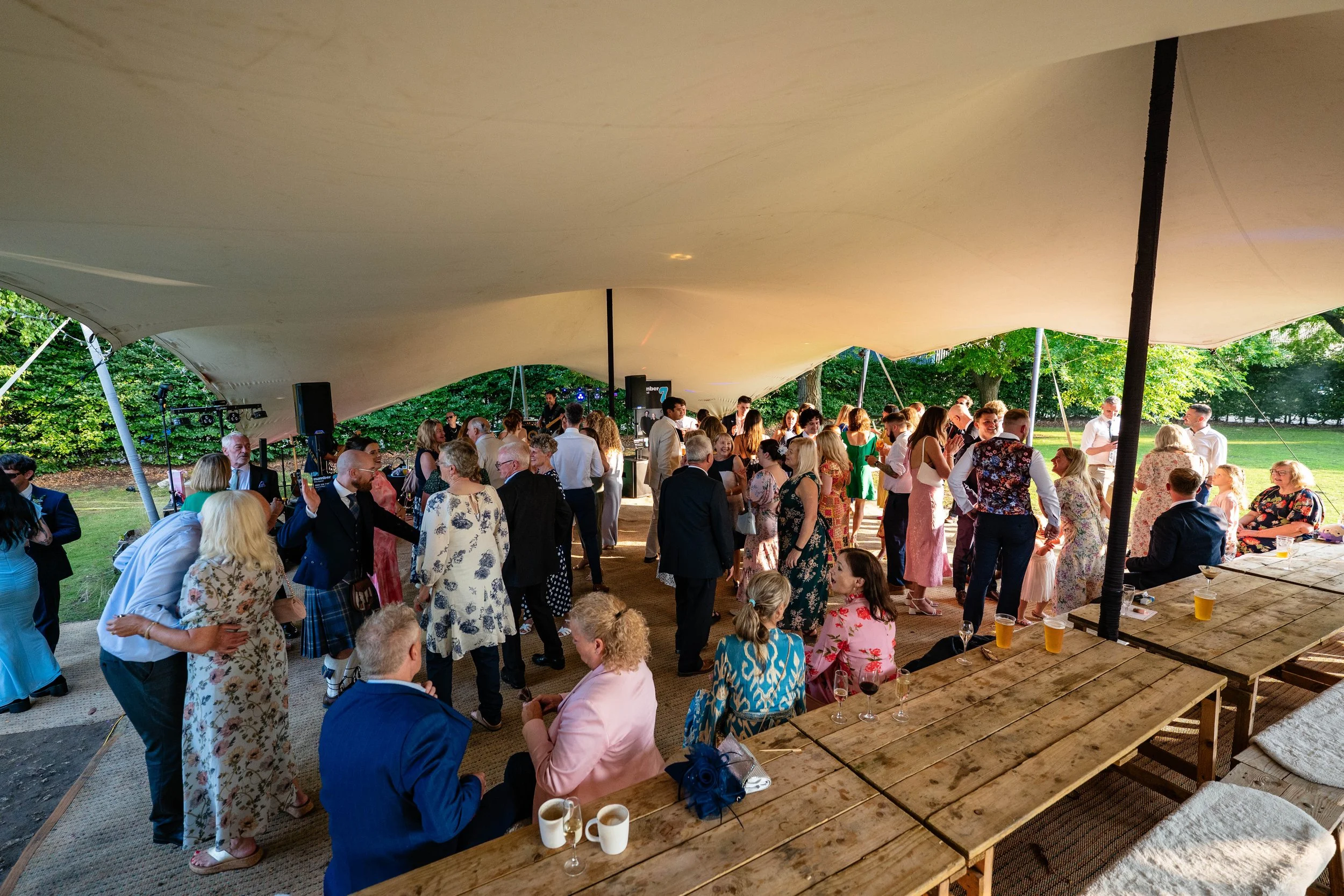 People gathering and dancing at an outdoor event under a large canopy, with greenery in the background.