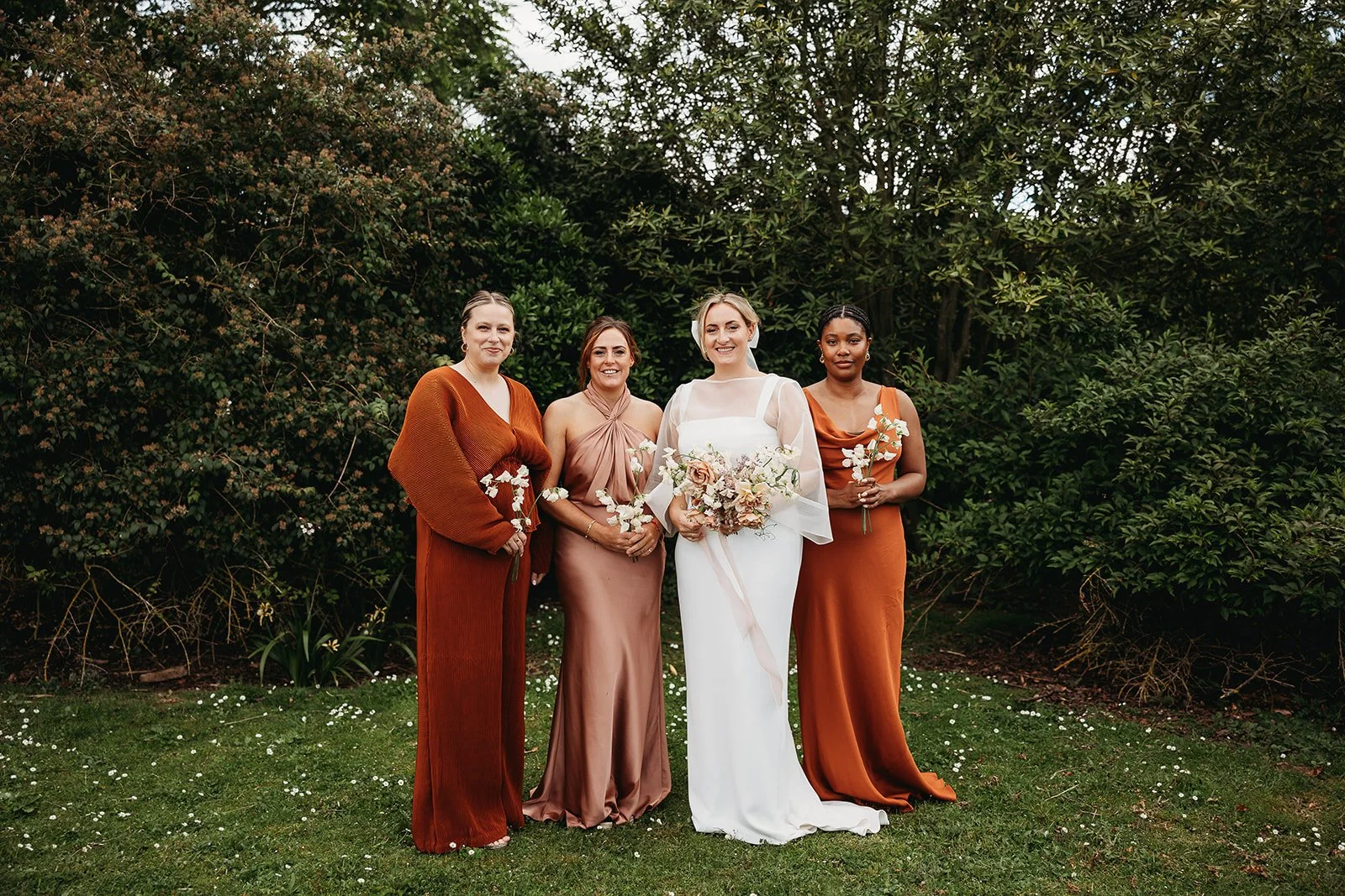 Four women, including a bride in a white wedding dress holding a bouquet, standing outdoors on grass with green bushes and trees in the background. The other women are dressed in elegant, rust-colored gowns and are holding small floral arrangements.