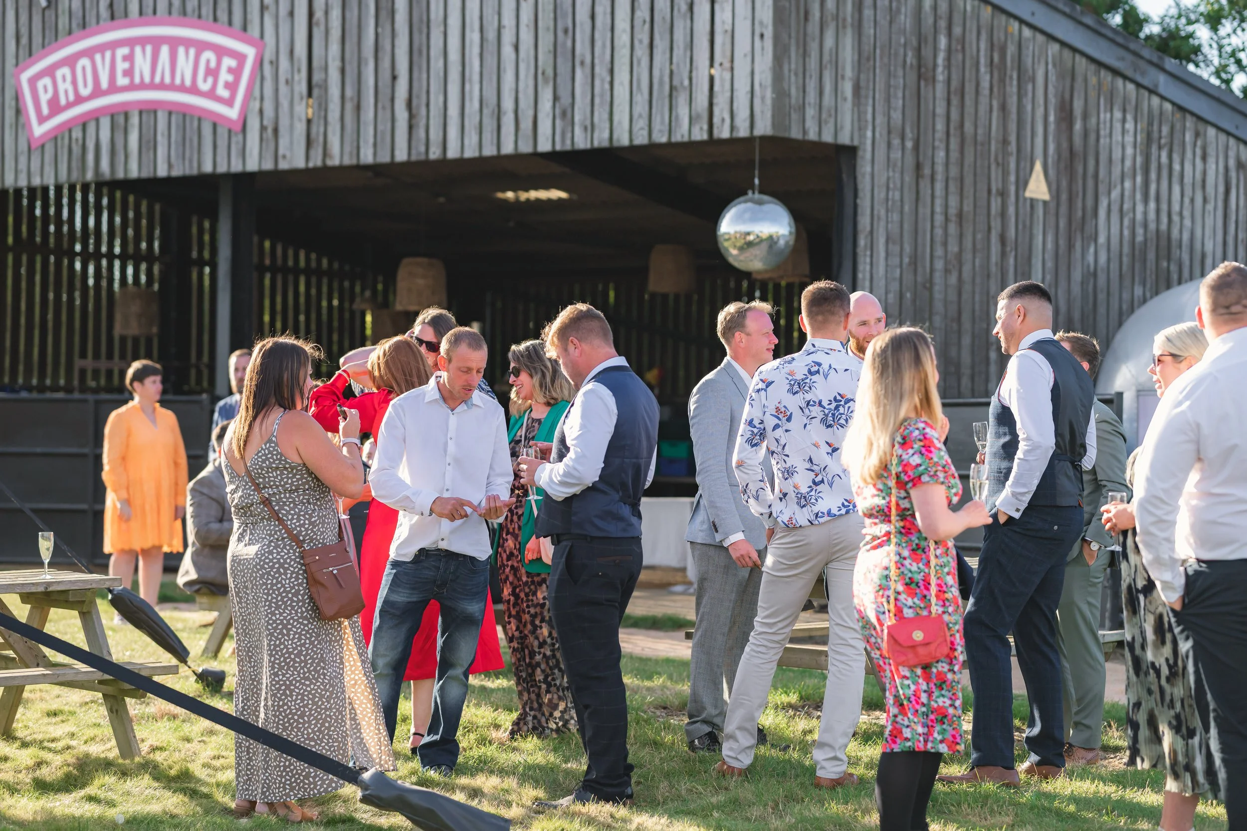 Group of people at an outdoor social gathering in front of a wooden barn with a pink and white Provence sign. Some guests are talking, holding drinks, and wearing festive, colorful attire.