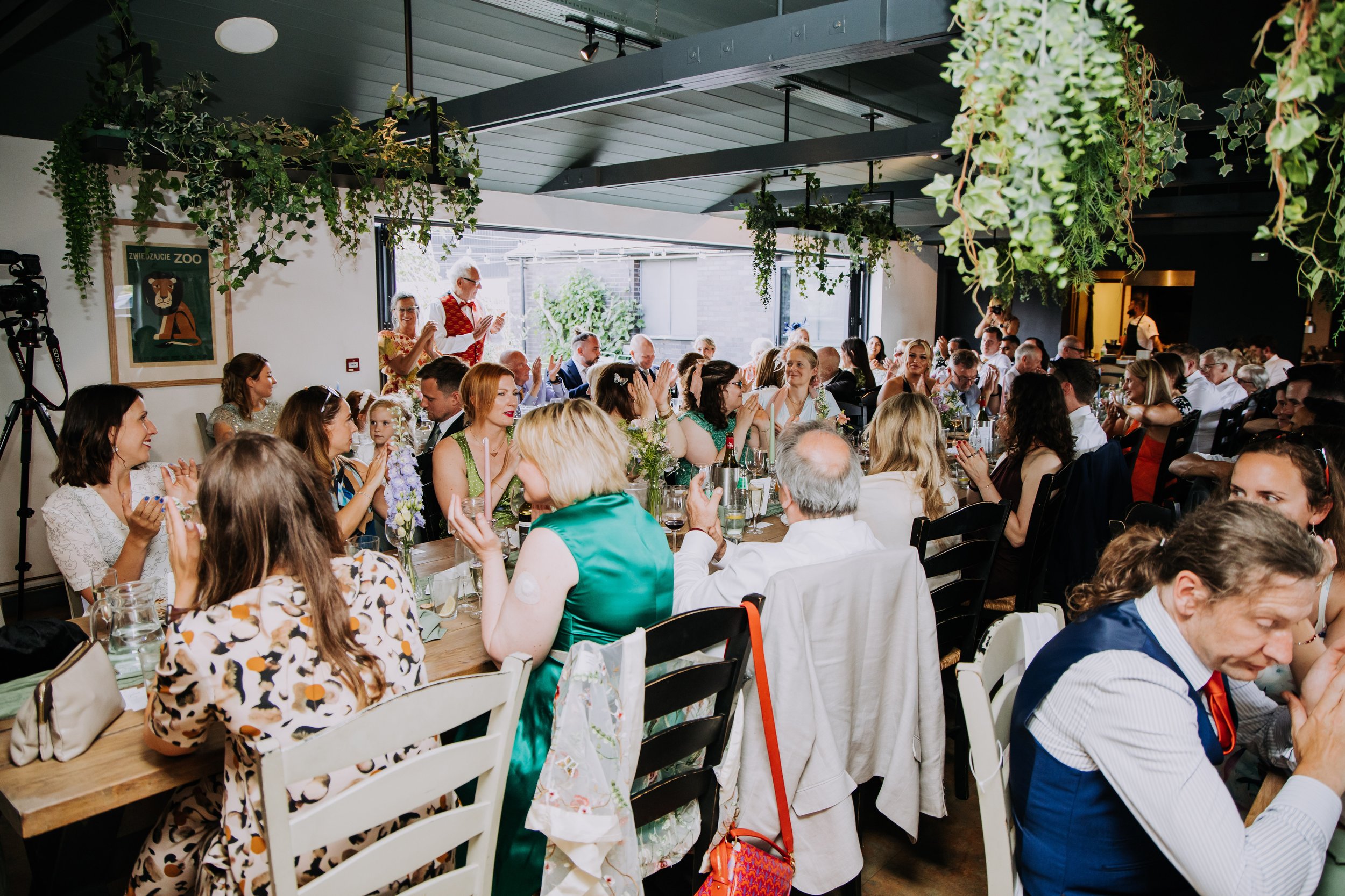 A large group of people gathered around tables in a decorated indoor venue, celebrating an event, with some standing and clapping, and others sitting and talking. The room is decorated with hanging plants and artwork, and there is natural light comin