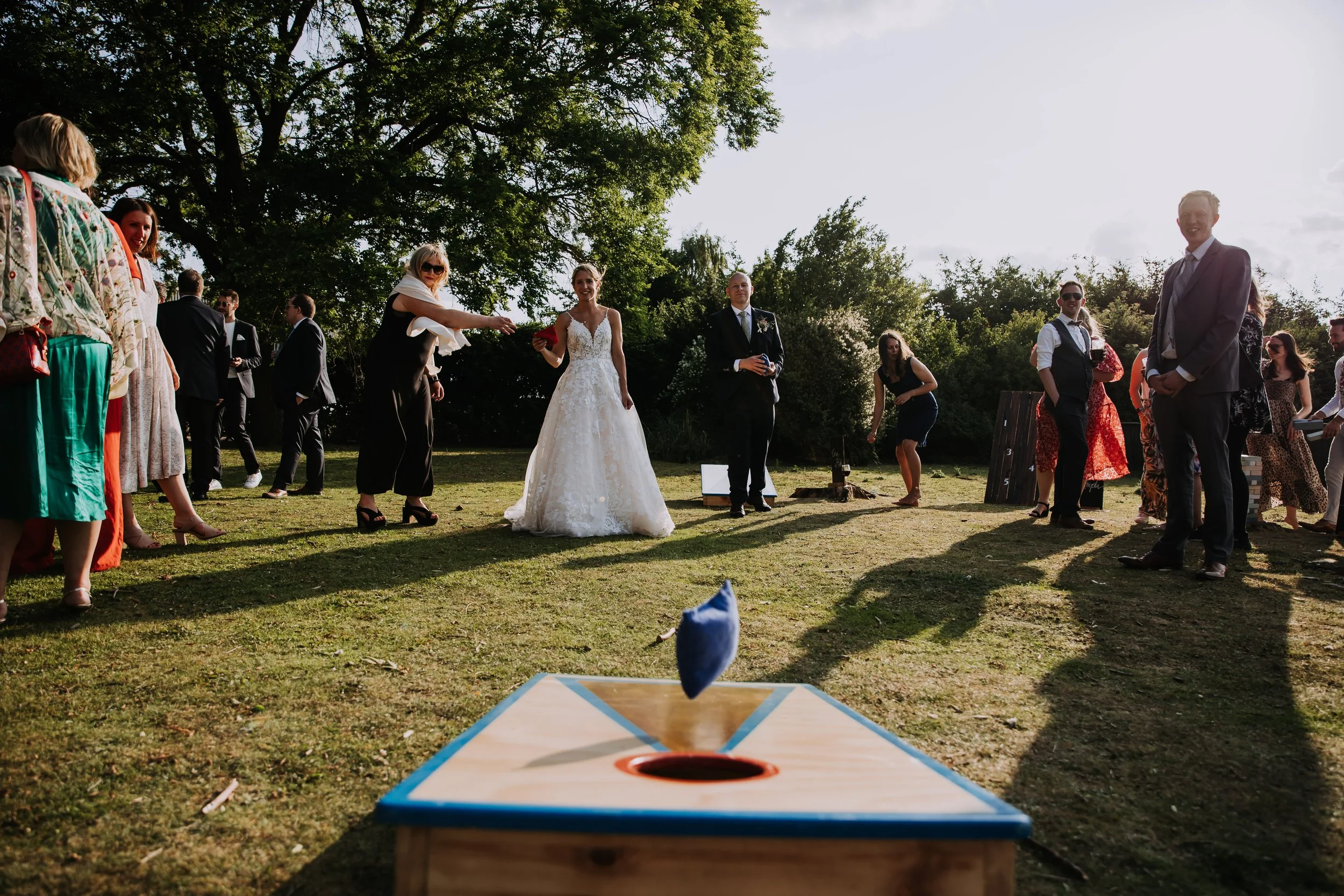 Wedding guests playing a game of cornhole outdoors with a bride in a white wedding gown and groom in a black suit in the background, surrounded by trees and sunlight.
