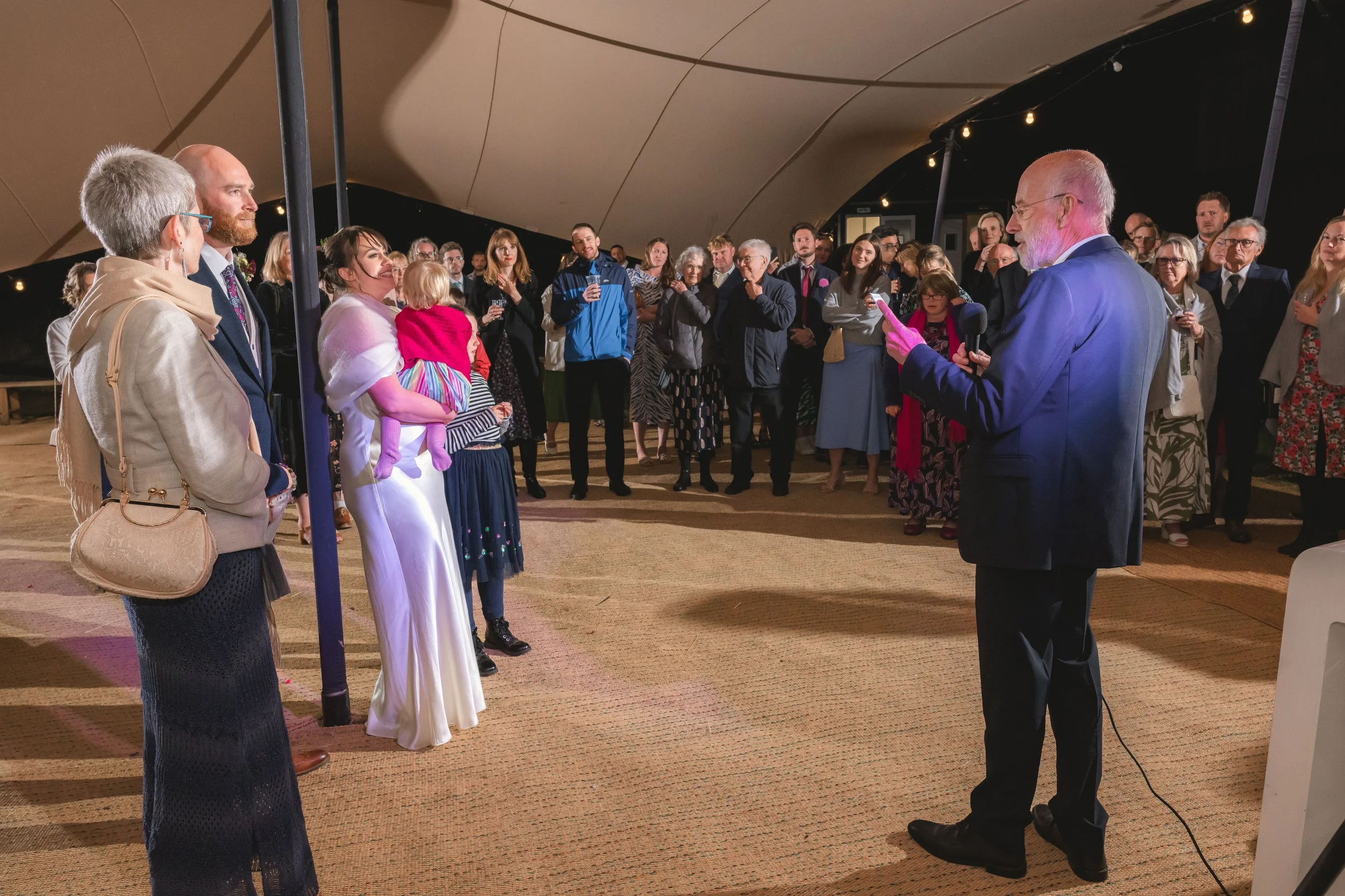 A man in a suit is speaking to a group of people gathered indoors under a large tent.