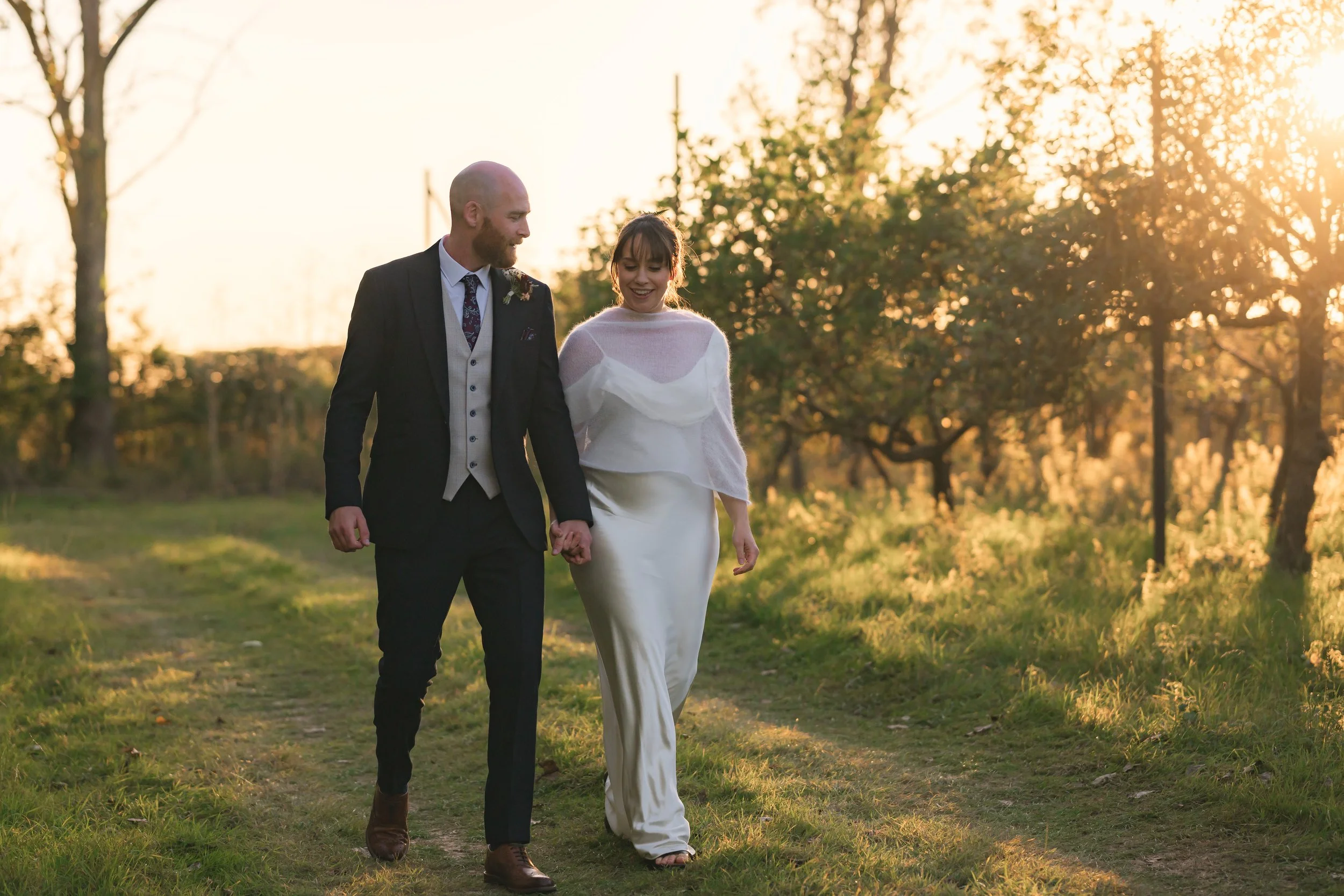 A bride and groom walking hand in hand outdoors during sunset, surrounded by trees and greenery.