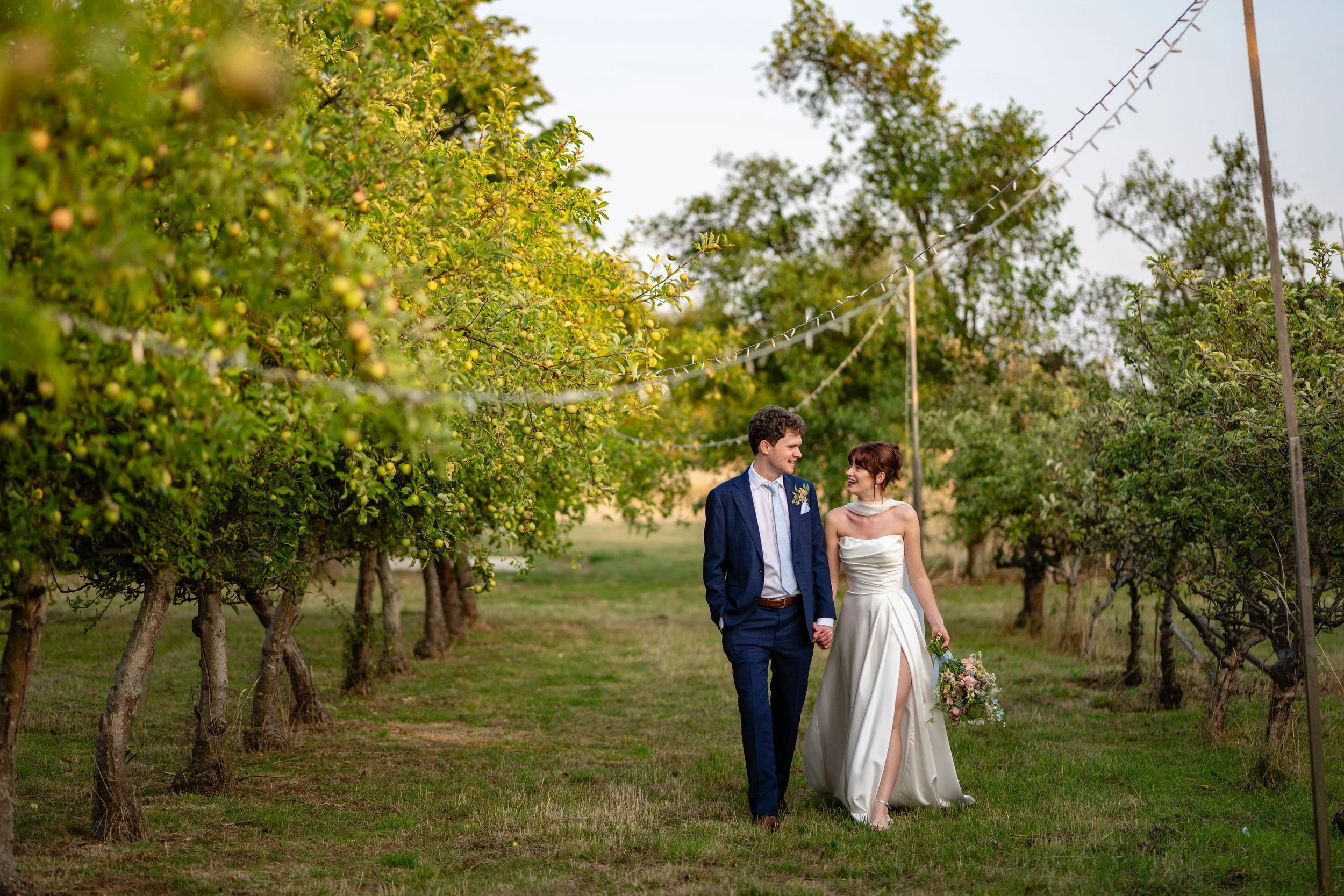 A bride and groom walking hand in hand through an orchard, surrounded by green trees, with the bride holding a bouquet of flowers and wearing a white gown, and the groom in a blue suit.