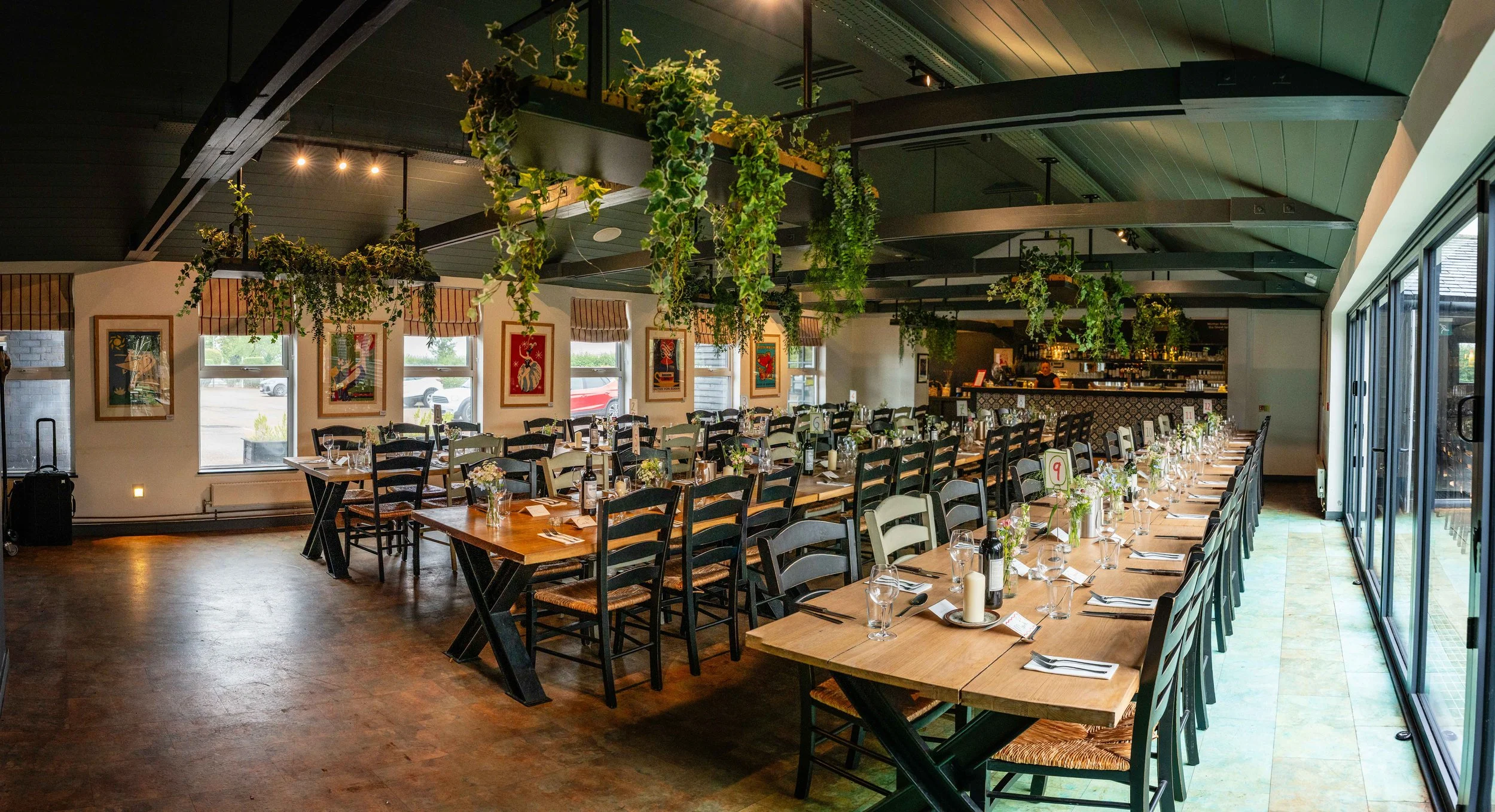 A restaurant interior with neatly set tables arranged for dining, decorated with flowers, wine bottles, and candles, with hanging greenery from the ceiling and framed artwork on the walls.