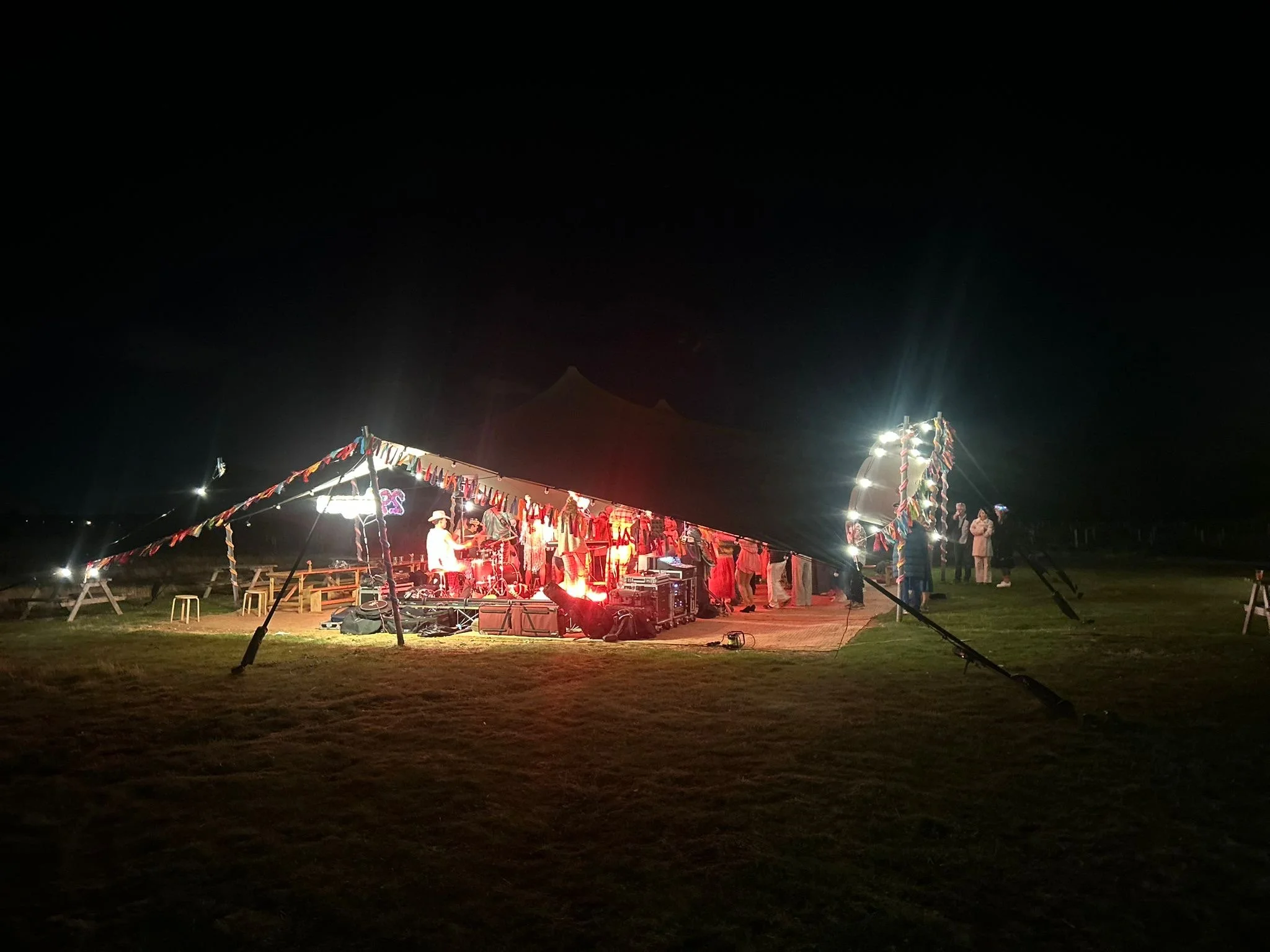 A nighttime outdoor music performance under a large tent decorated with colorful streamers, with people standing and sitting nearby, illuminated by stage lights.