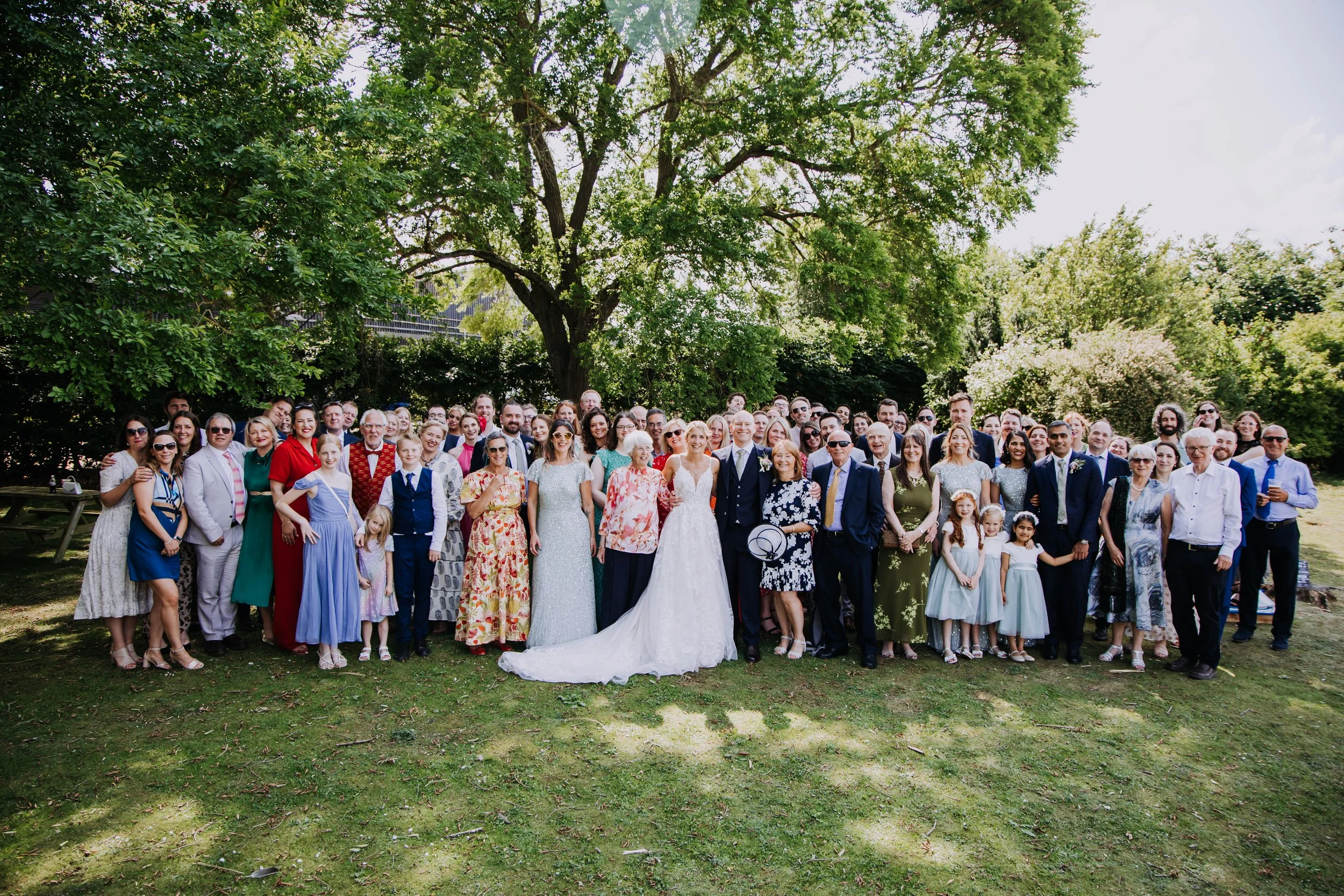 A large group of people gathered outdoors on a grassy area under a tree on a sunny day, celebrating a wedding with a bride and groom in the center.