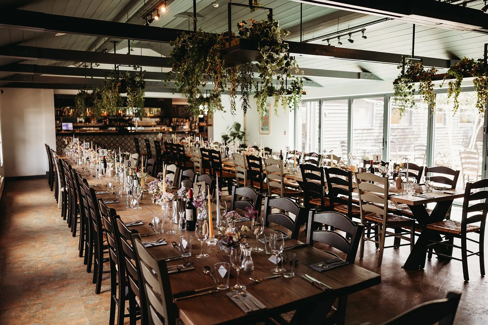 Indoor dining area with long wooden tables set for a formal event, decorated with floral centerpieces, candles, wine bottles, and glassware, with hanging greenery above and large windows letting in natural light.