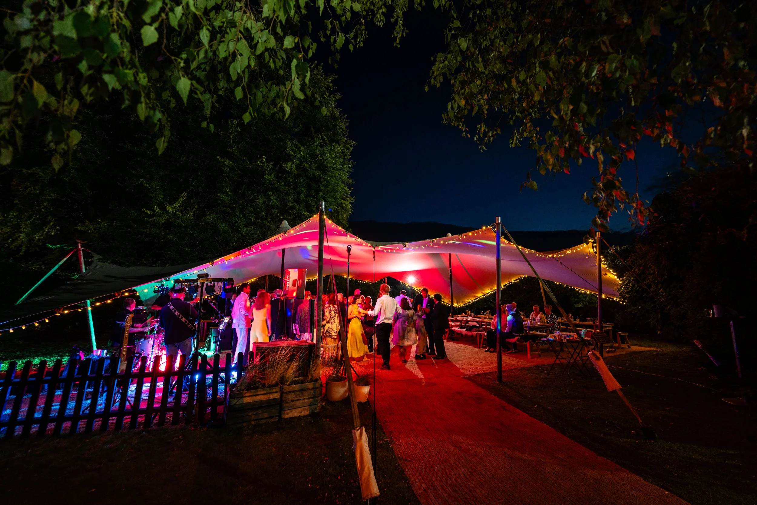 Nighttime outdoor party under a large tent with colorful lighting, guests dancing and socializing, surrounded by trees.