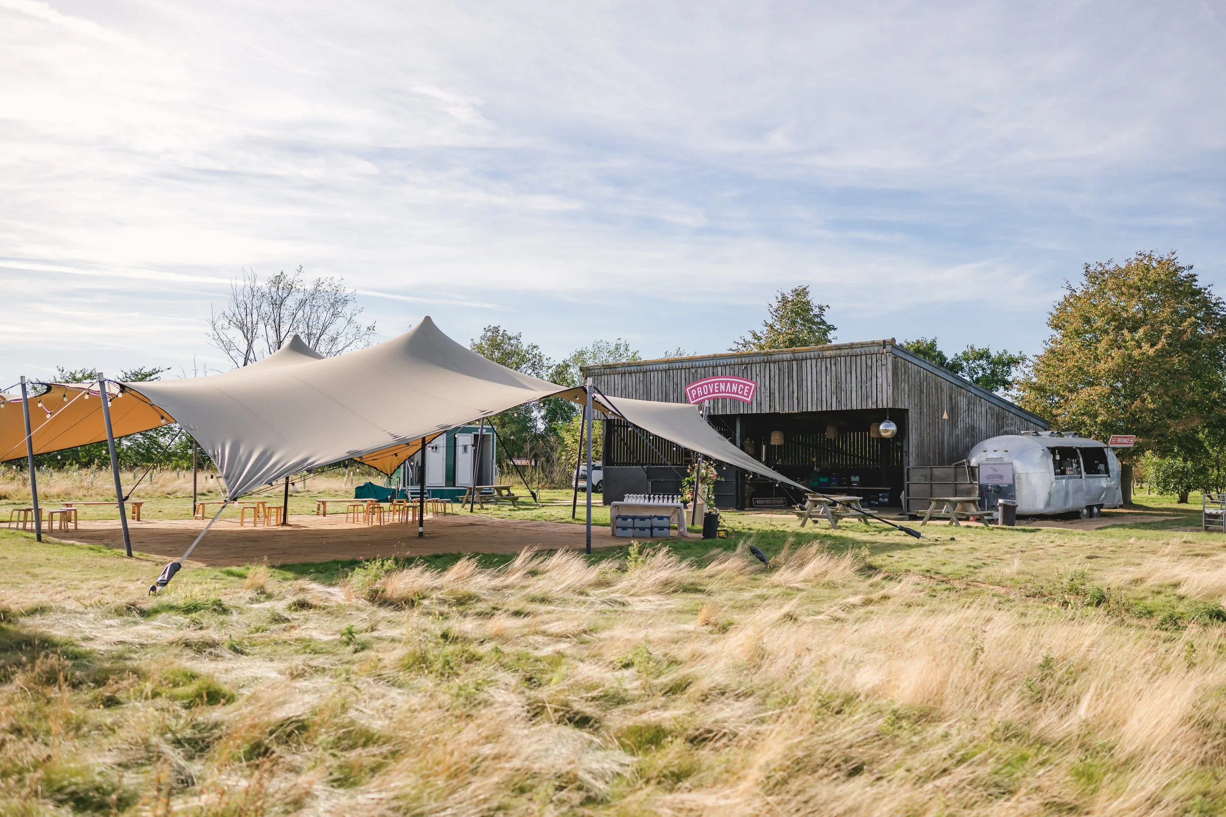 Outdoor scene with a wooden building labeled 'Provenance,' a silver Airstream trailer, and a large white tent with support poles, set in a grassy area with trees and a clear sky.