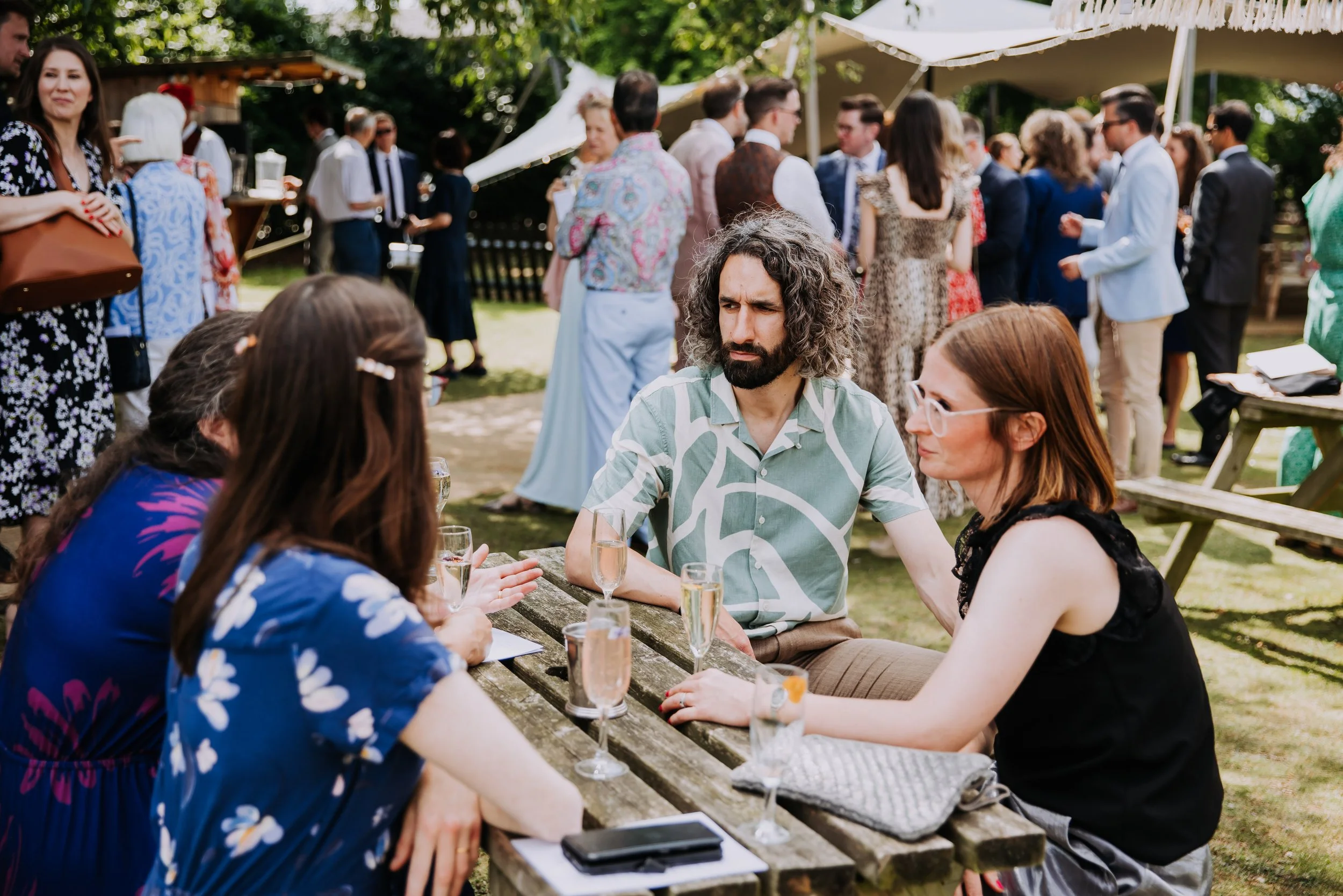 A group of people socializing at an outdoor party, with some standing and chatting under a canopy, while three women and one man sit at a rustic wooden picnic table in the foreground, engaged in conversation and drinking champagne.
