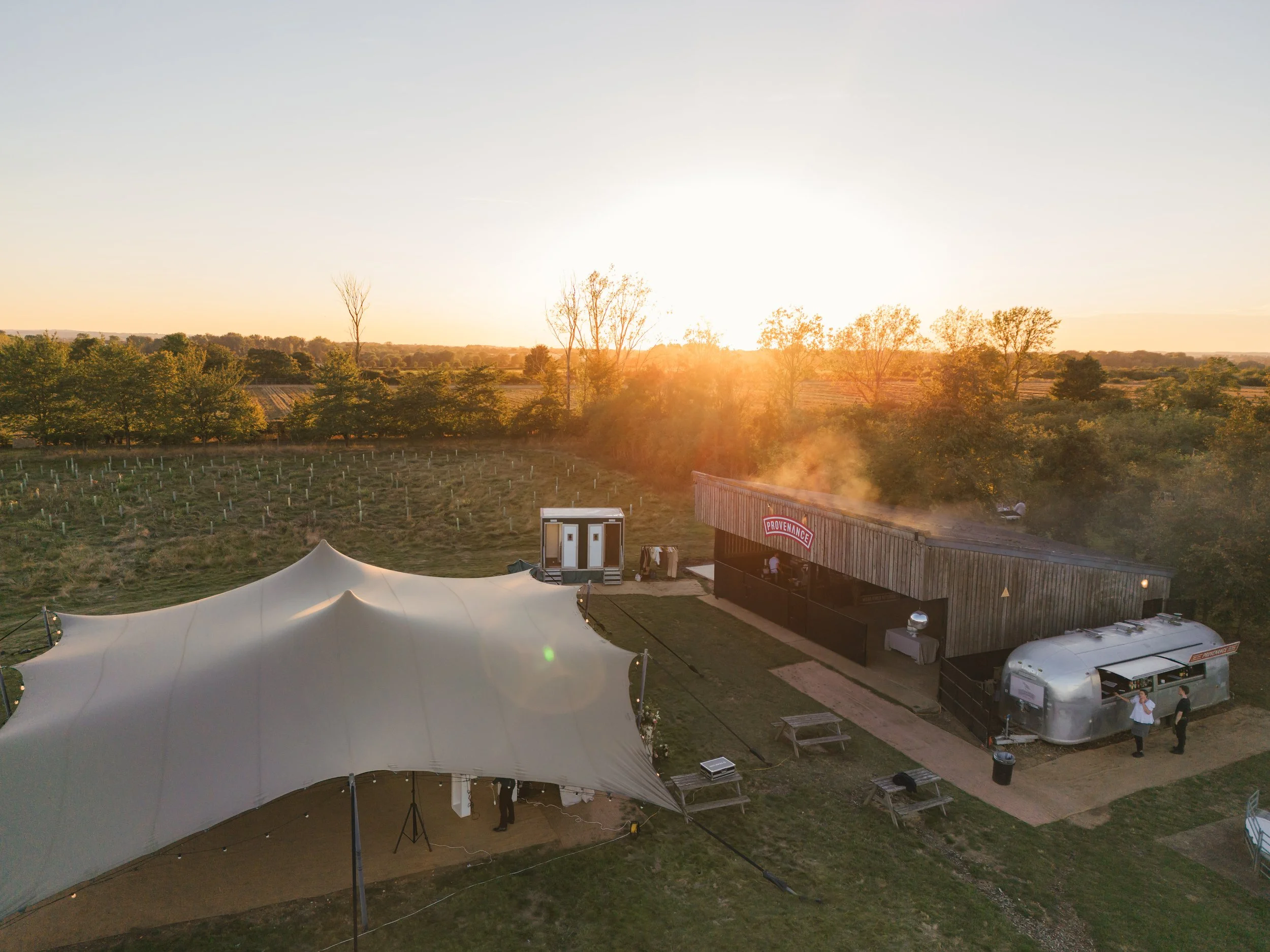 Aerial view of a farm or winery at sunset, featuring a large white tent, a wooden building with a sign that reads 'Provenance,' a silver Airstream trailer, and picnic tables.