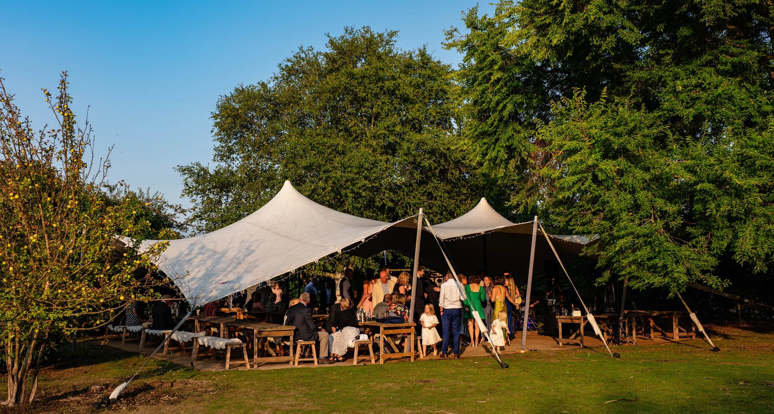 Outdoor gathering of people under a large white tent with wooden benches and tables, surrounded by green trees on a sunny day.