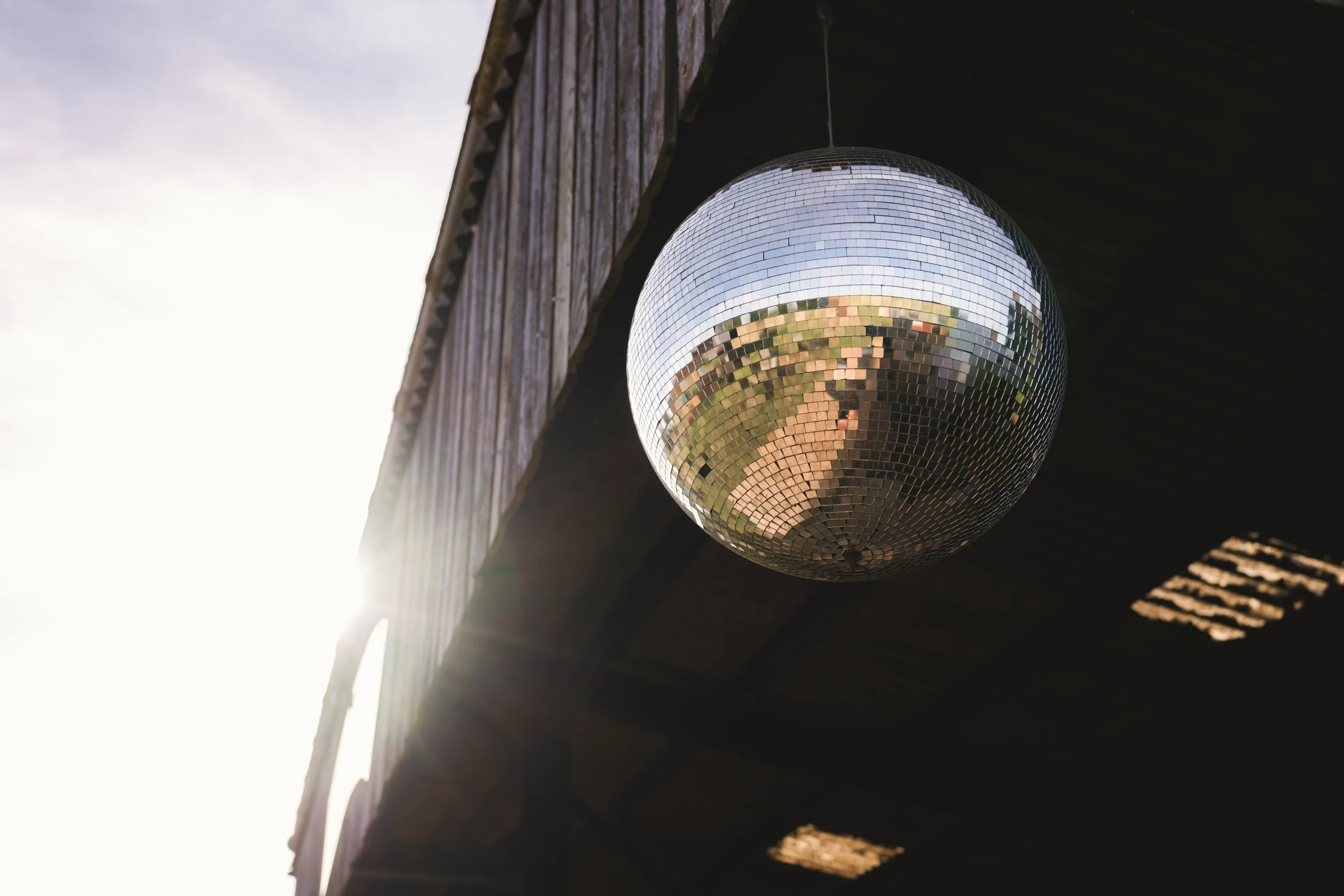 A disco ball hanging under a dark wooden ceiling, reflecting the outdoor scenery including the sky and trees.