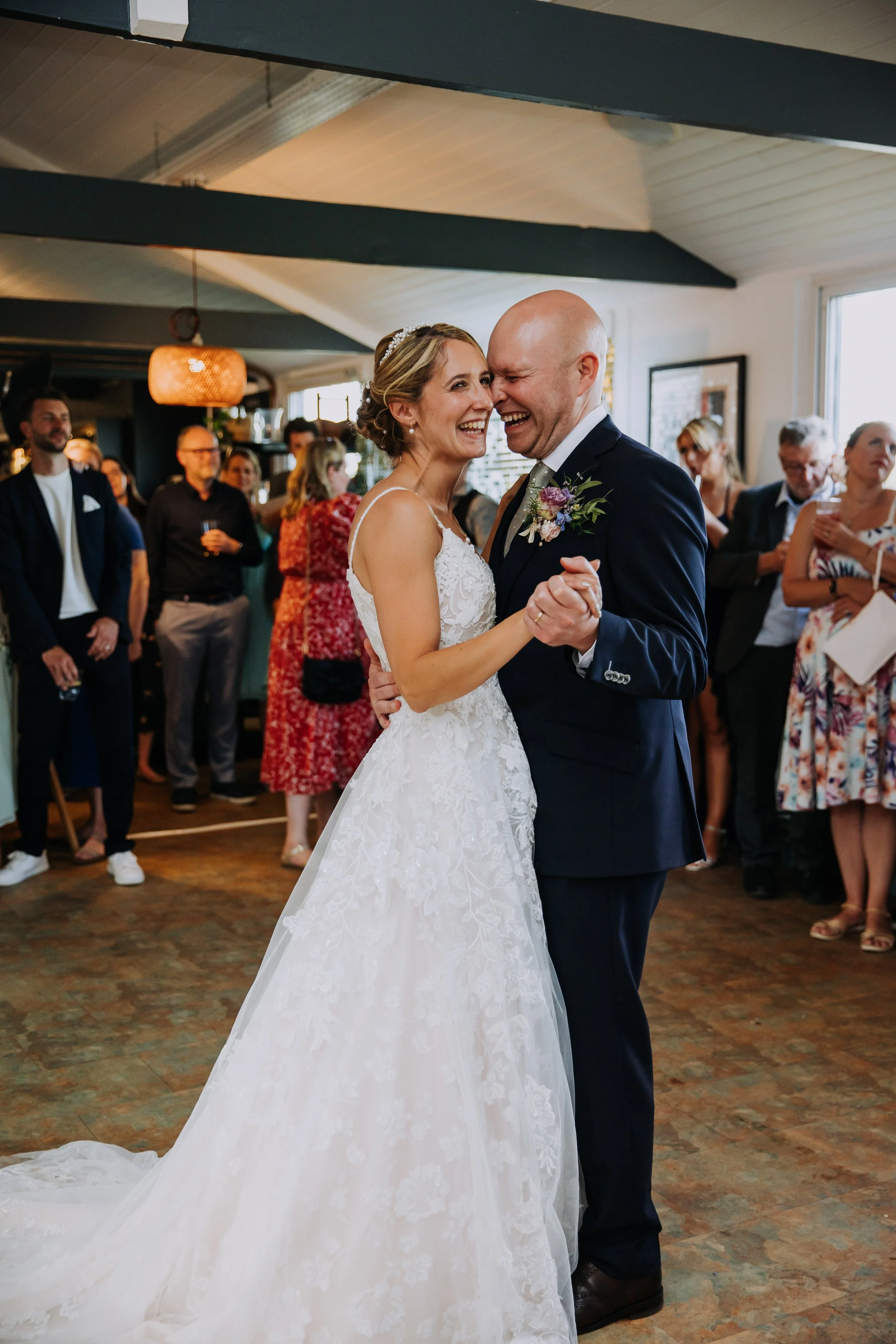 Bride and groom dancing at wedding reception, smiling and holding hands, with guests watching in the background.