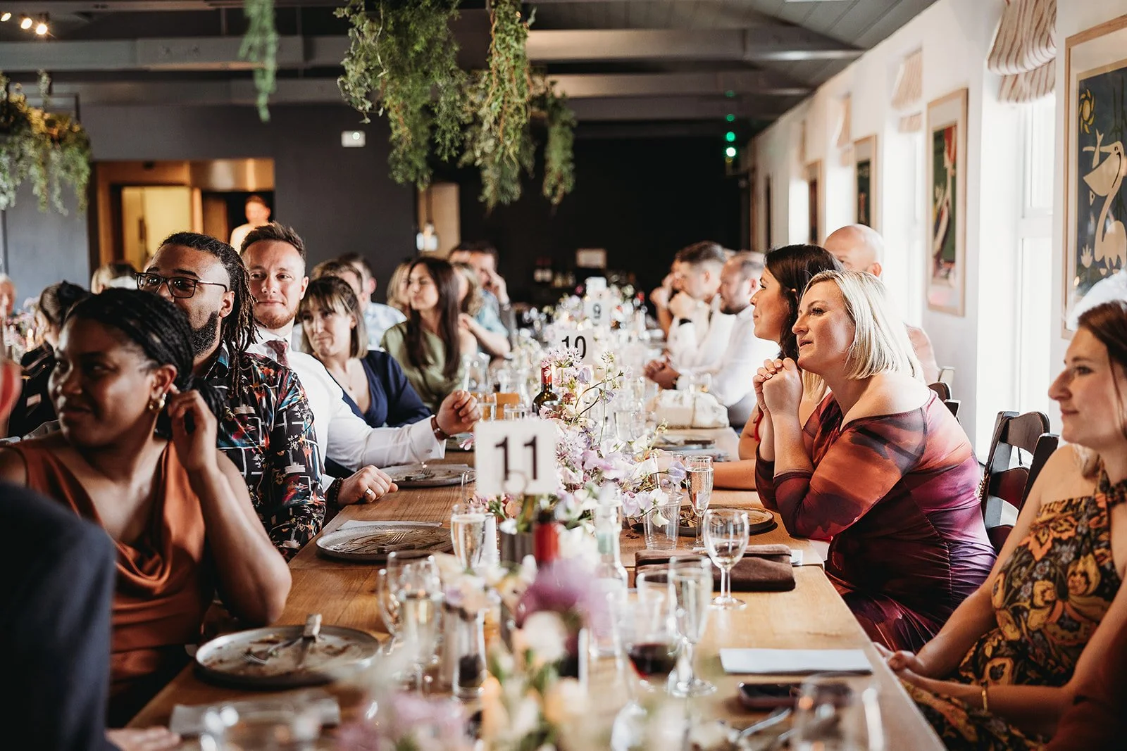 A diverse group of people sitting at a long dining table during a celebration or formal event in a decorated room with floral arrangements and artwork on the walls.