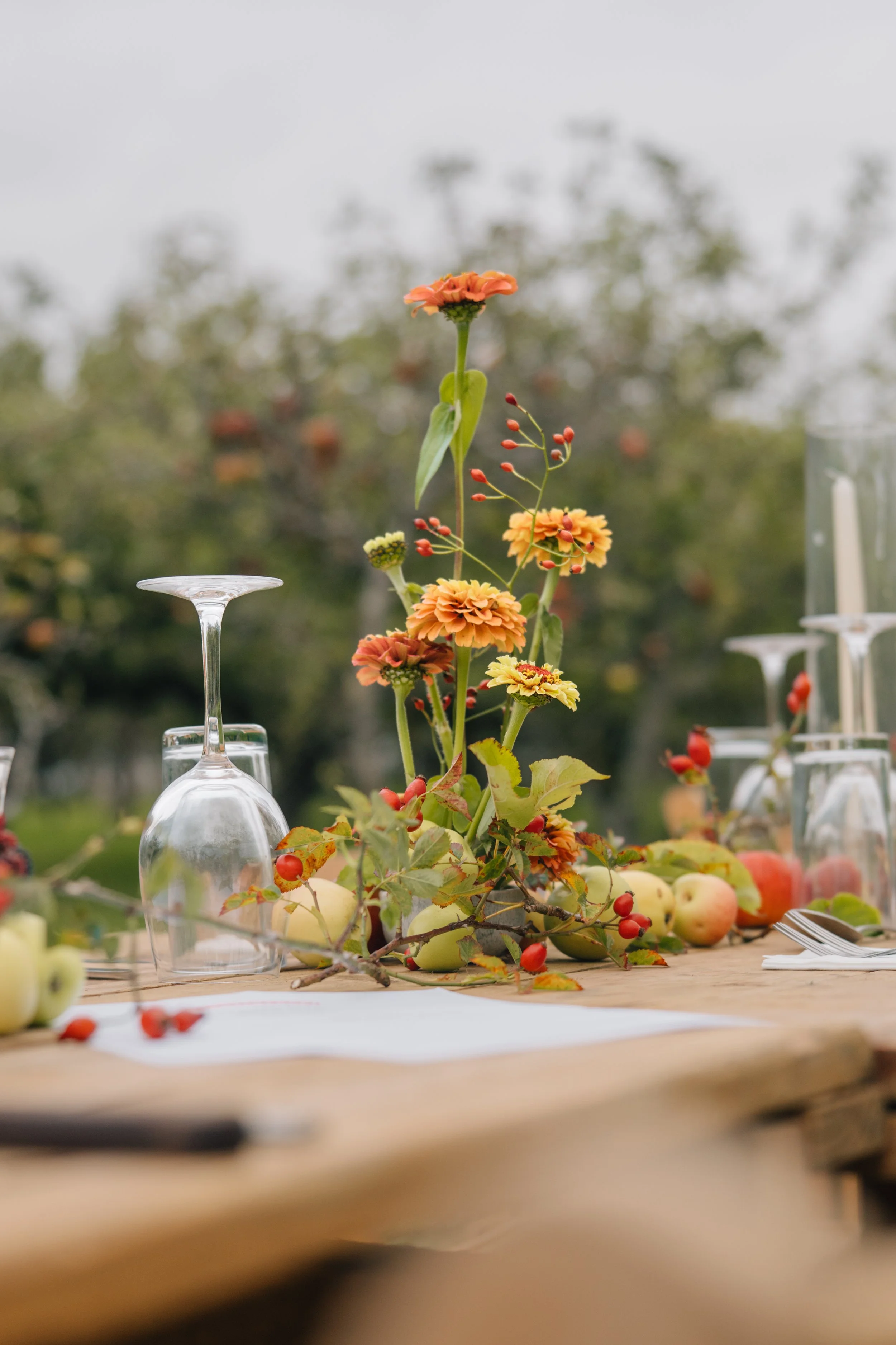 Table centerpiece featuring orange and yellow flowers, apples, and berry branches, with upside-down wine glasses on a wooden table outdoors.