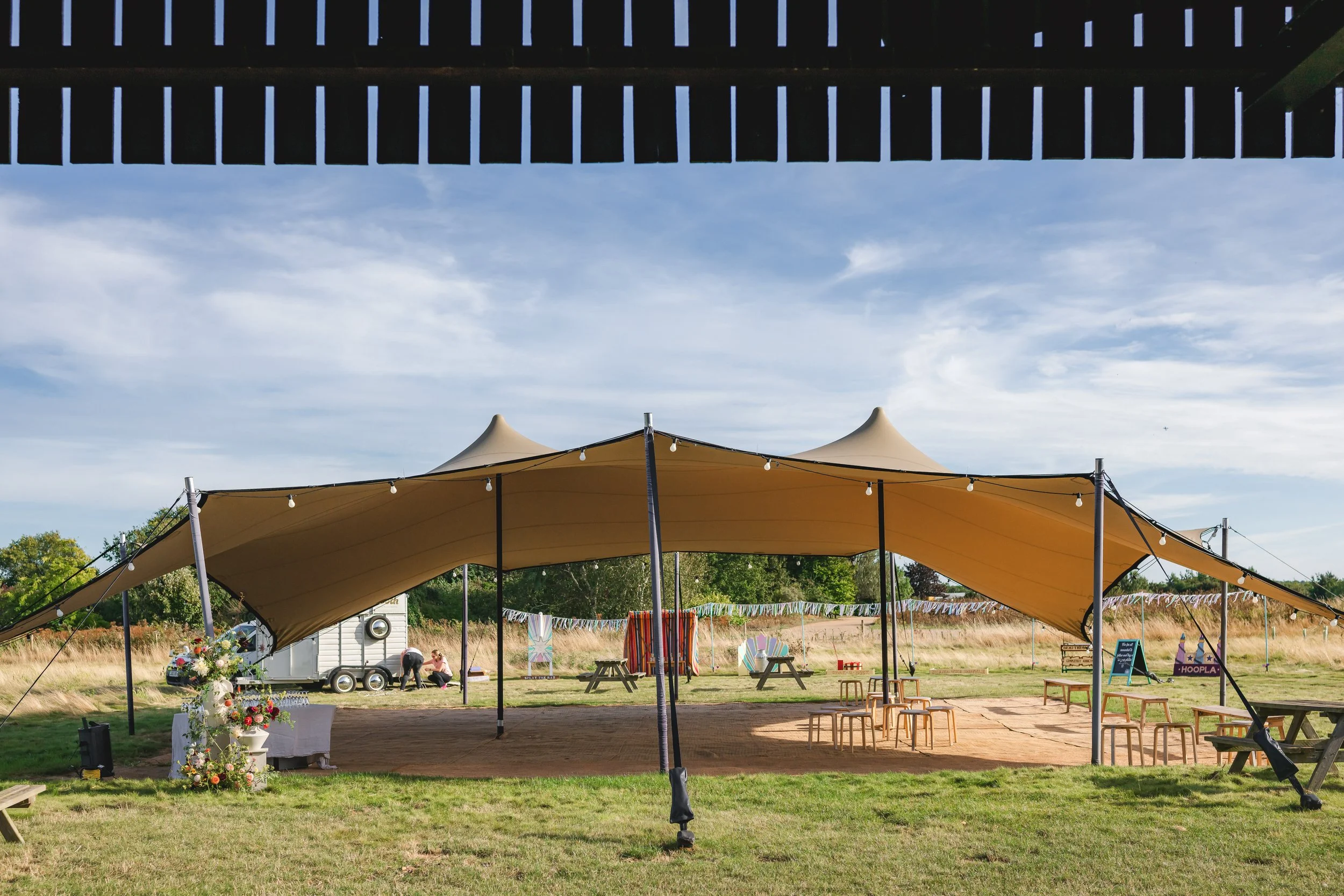 Outdoor event space with a large beige canopy tent decorated with string lights, surrounded by benches and tables, set in a grassy field with a wooden stage, colorful flags, and a clear blue sky with some clouds.