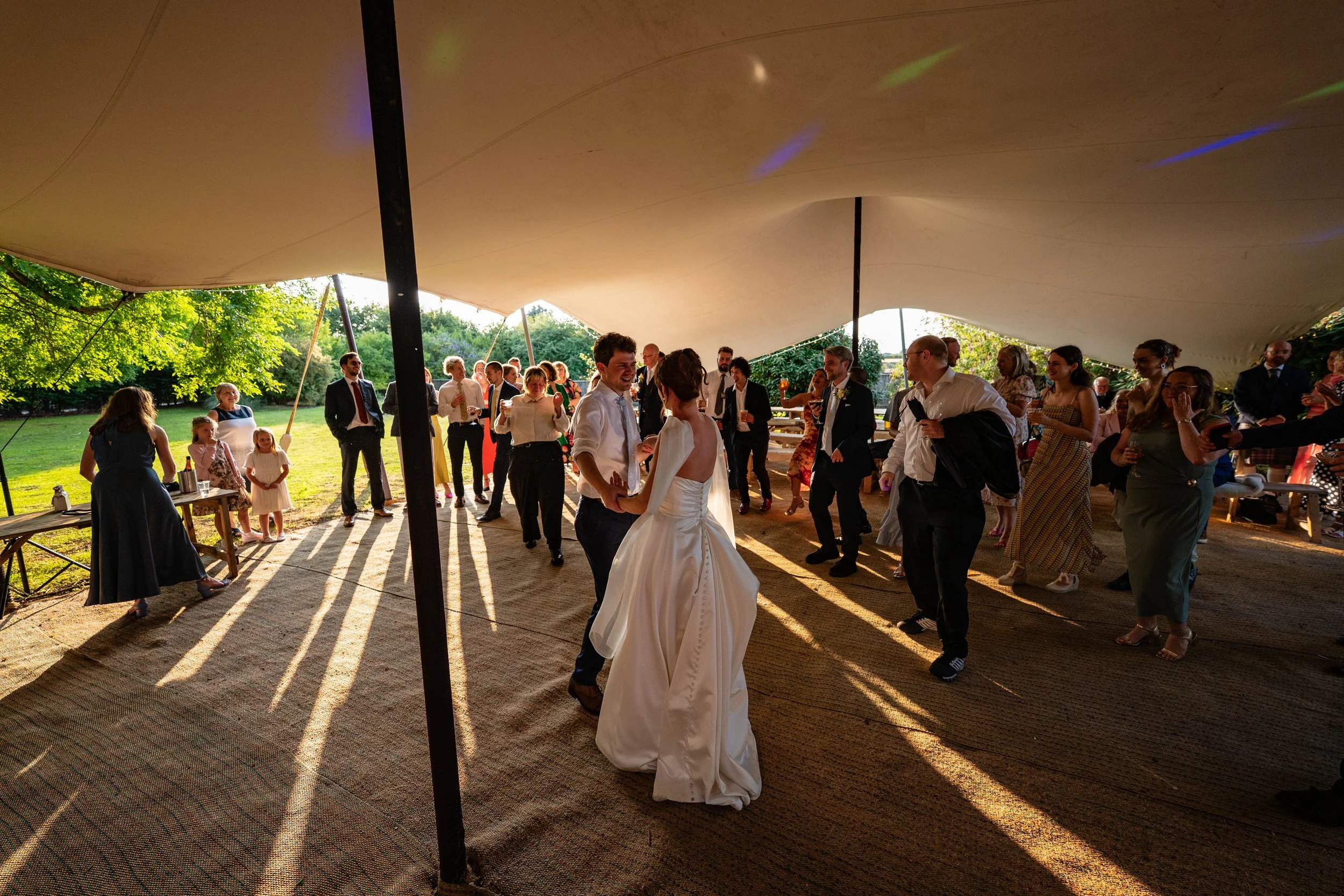 People dancing under a large tent at an outdoor event, likely a wedding reception, with the sun casting long shadows.
