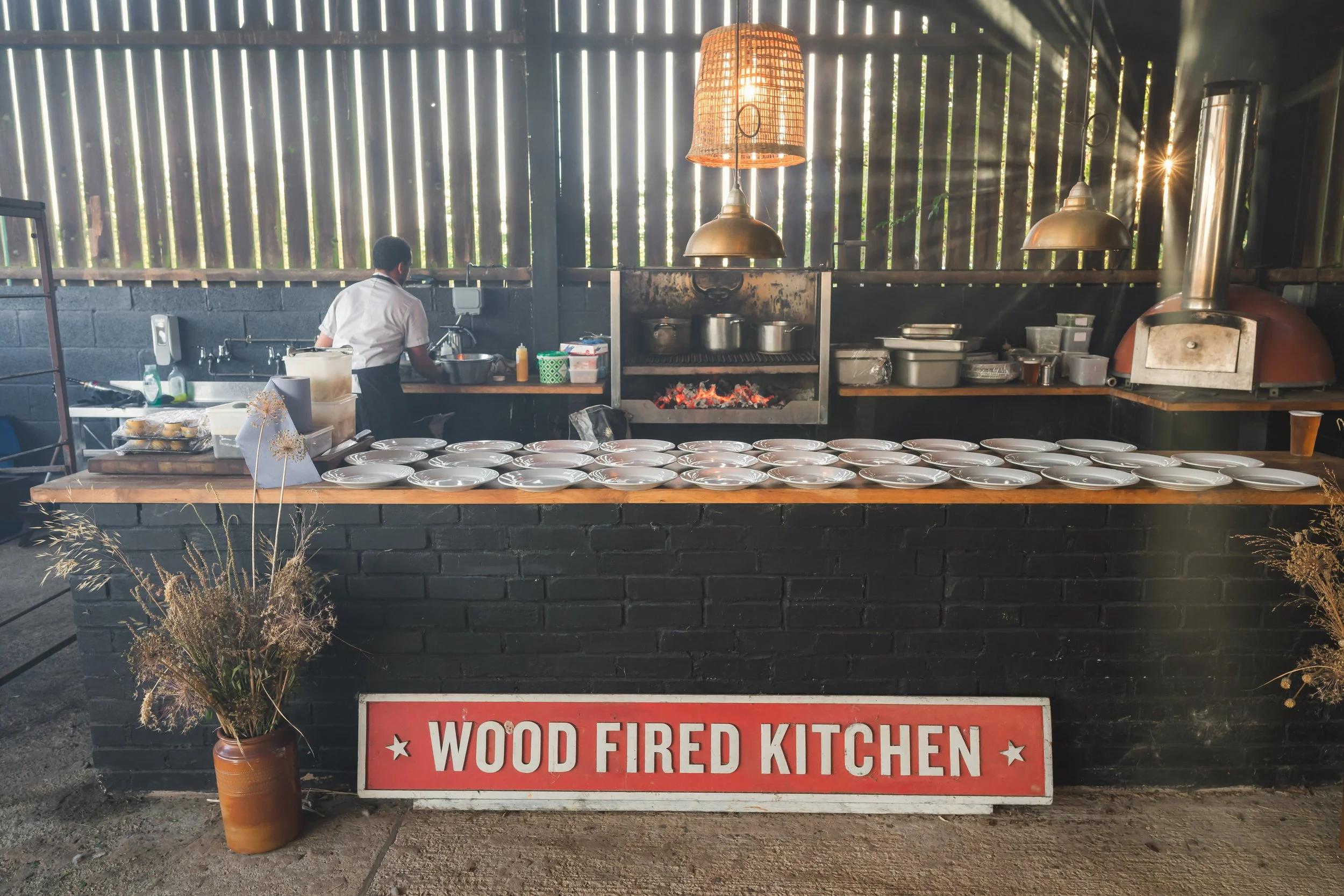 An outdoor kitchen with a brick counter and a large wood-fired oven. Plates are arranged on the counter, and a person is cooking in the background. There is a sign that reads 'Wood Fired Kitchen' on the ground in front of the counter. Decorative drie