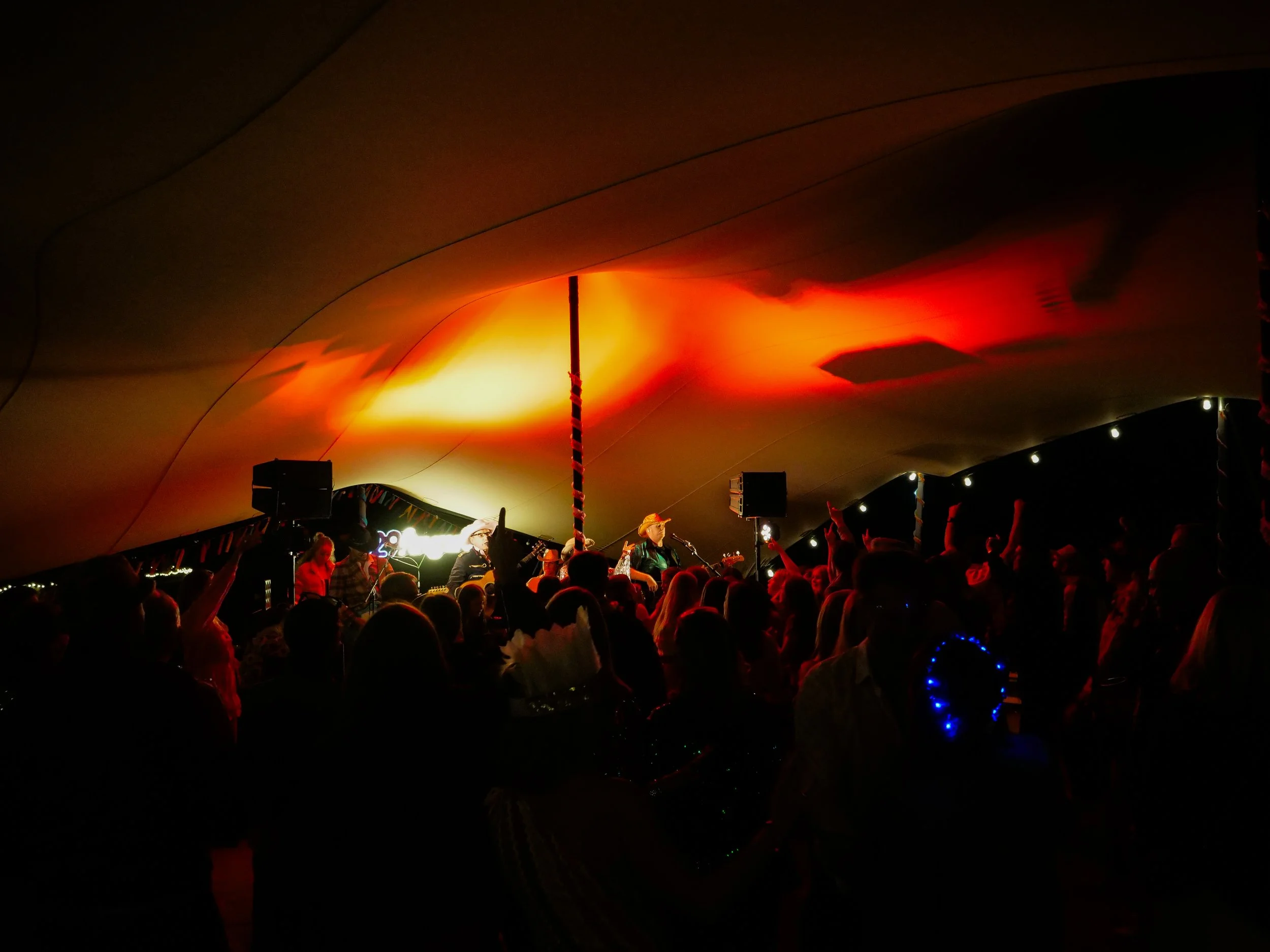 Musicians perform on stage under a curved ceiling illuminated with orange and yellow lights, while a crowd dances and watches.