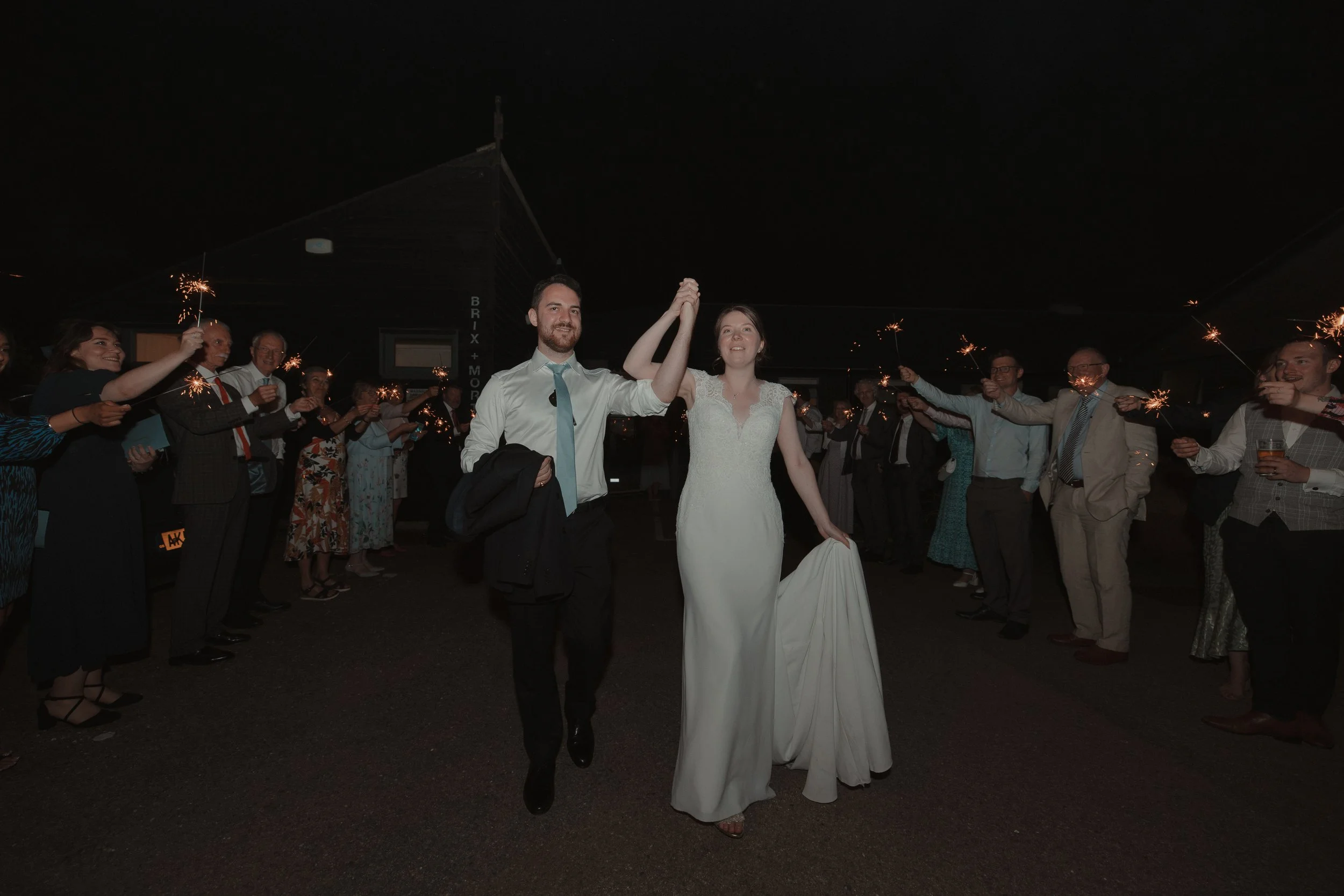 A newlywed couple holding hands and smiling as they walk through a line of guests holding sparklers at night during a wedding celebration.