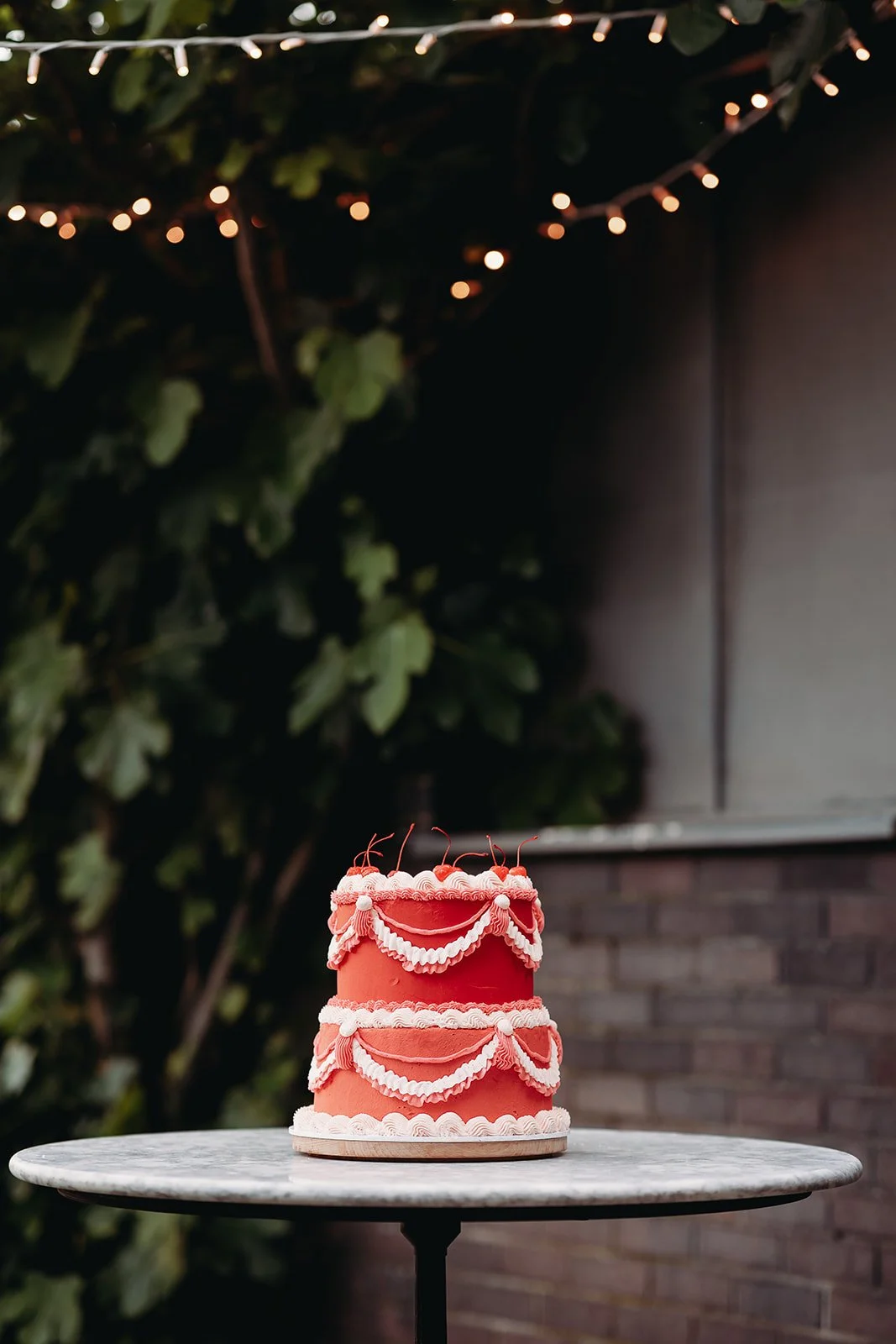 Two-tiered pink birthday cake with white icing decorations on a marble table, illuminated by string lights in an outdoor setting with dark background and greenery.