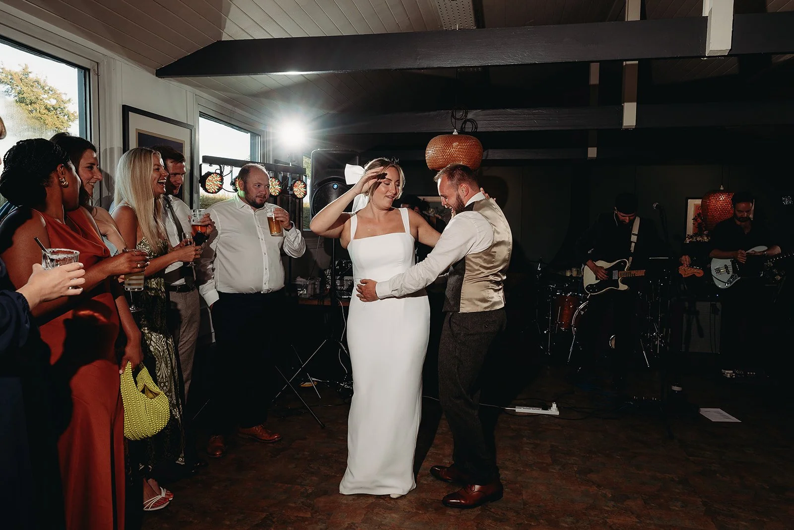 A bride and groom dance together at their wedding reception, surrounded by guests holding drinks, with a live band playing in the background.