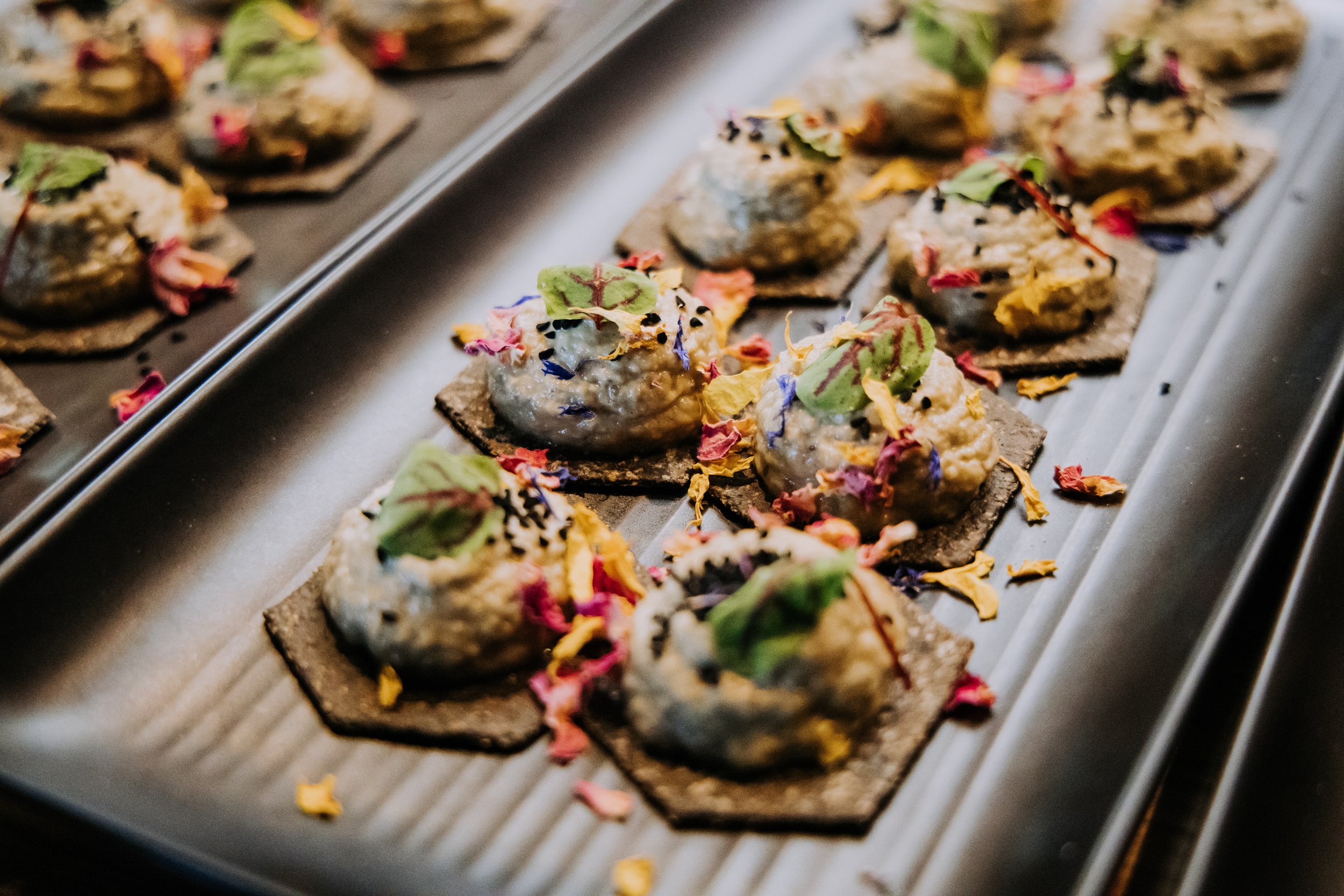 Appetizers with creamy topping on crispy crackers, garnished with edible flowers and herbs on a metal tray.