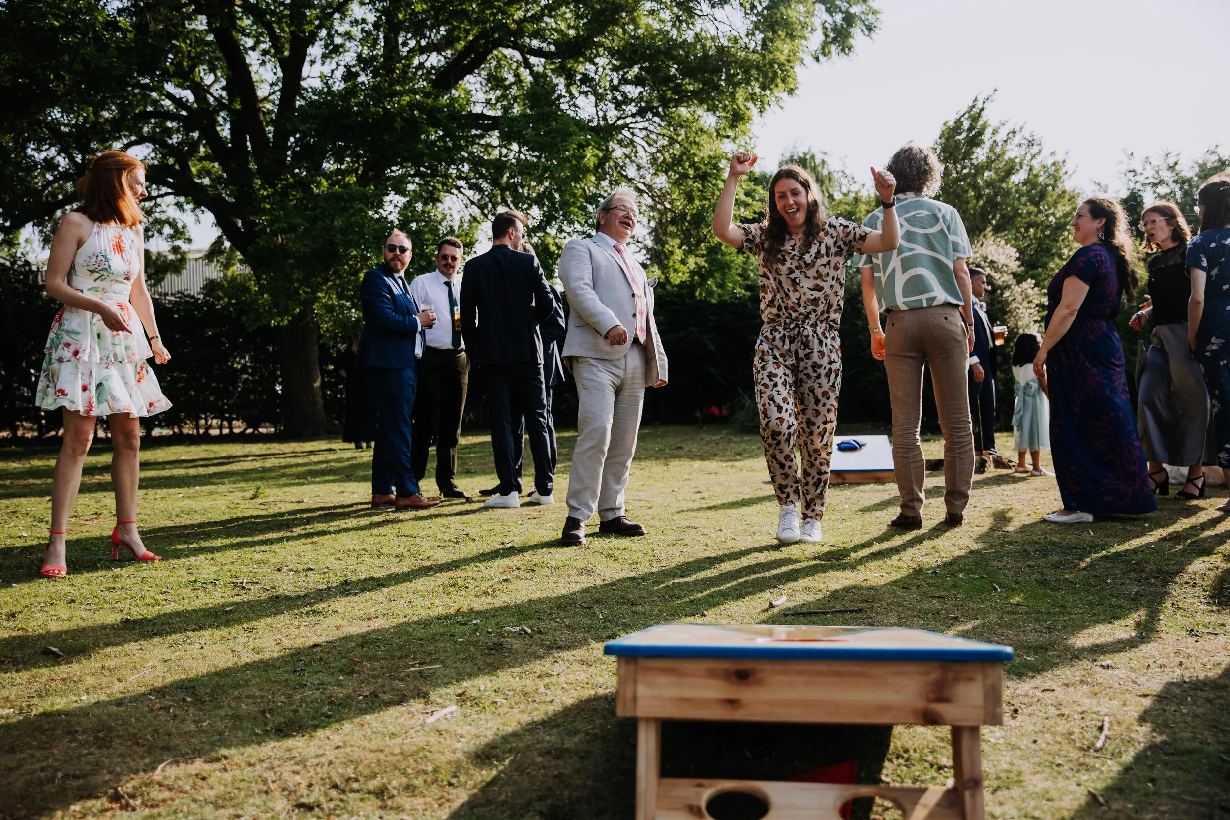 People gathered outdoors on a sunny day, some playing a game of cornhole, with trees and sunlight in the background.
