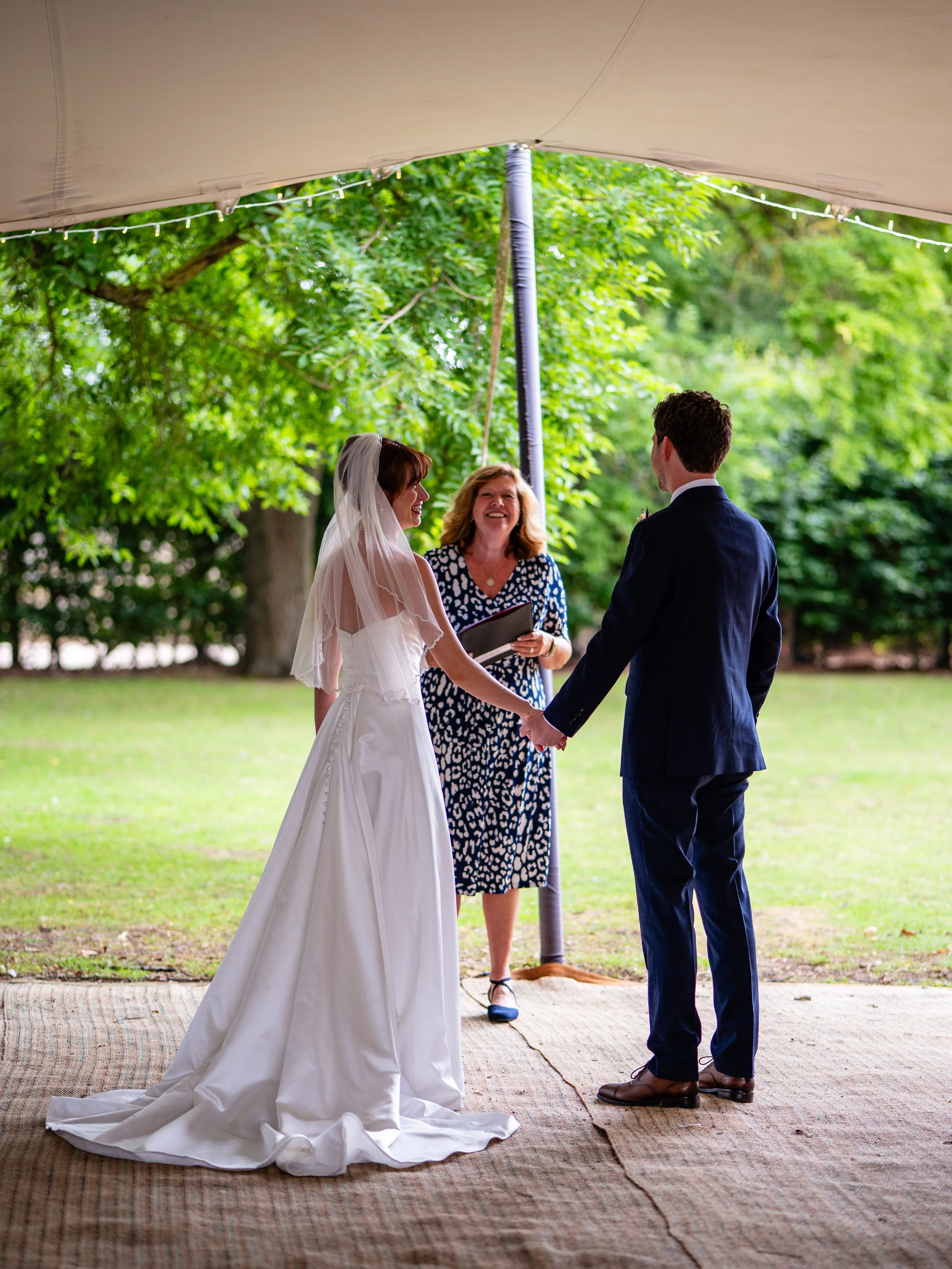 A bride and groom hold hands during an outdoor wedding ceremony under a tent, with a woman officiant smiling in the background, surrounded by greenery.