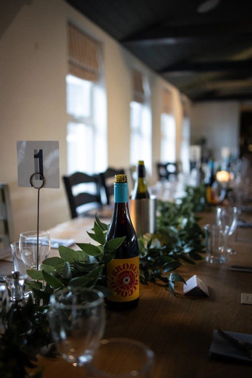 A long dining table decorated with greenery, wine bottles, empty glasses, and table number 1 card in a warmly lit room with windows and chairs.