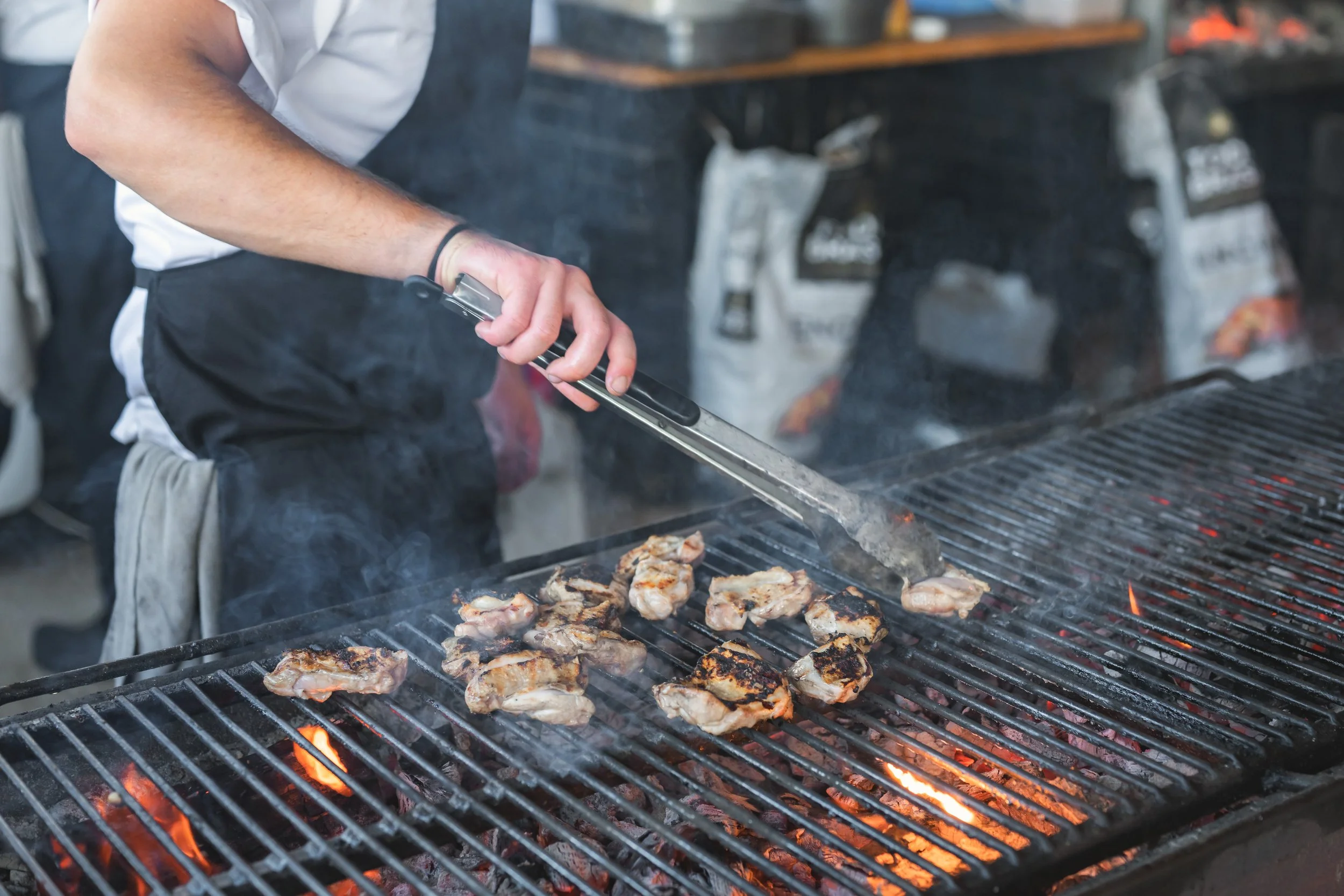Person grilling chicken pieces on a barbecue, using tongs, with flame and smoke visible.