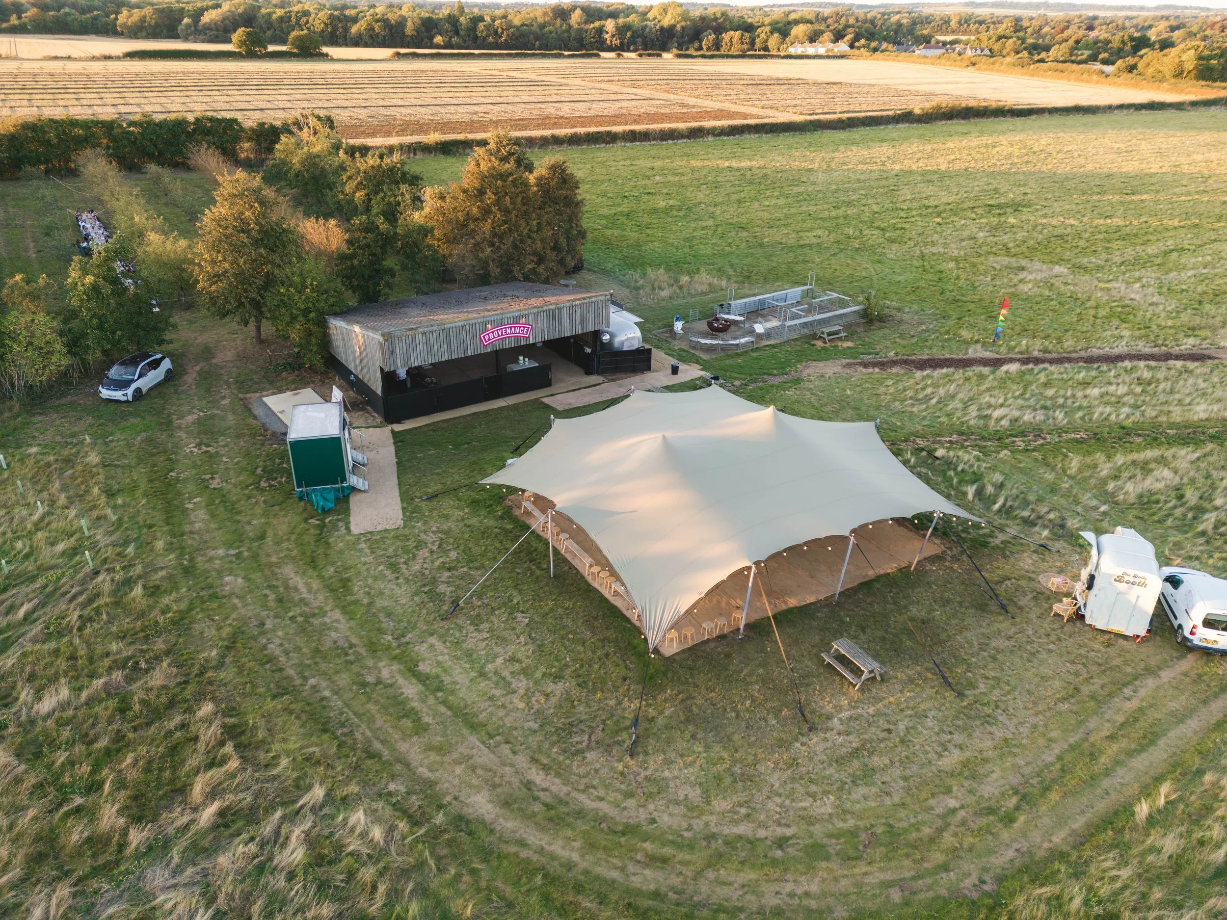 Aerial view of an outdoor event setup in a rural field featuring a large beige canopy tent, a small stage area with a black backdrop, and surrounding vehicles including a food truck, with trees and farmland in the background.