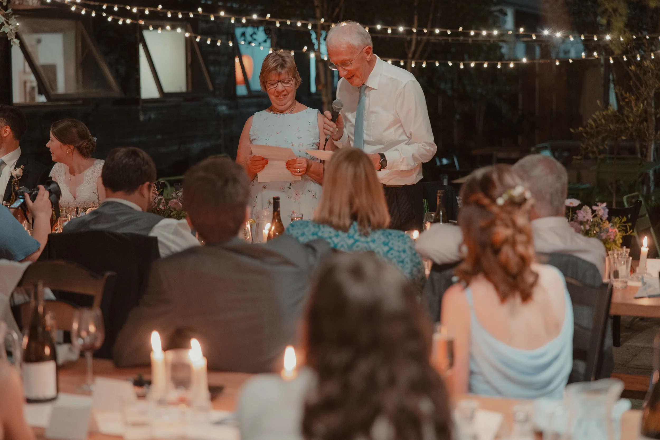 At a wedding reception, an older man and woman are standing and reading vows or a speech, surrounded by seated guests at decorated tables with candles and flowers, under string lights and a dark outdoor setting.